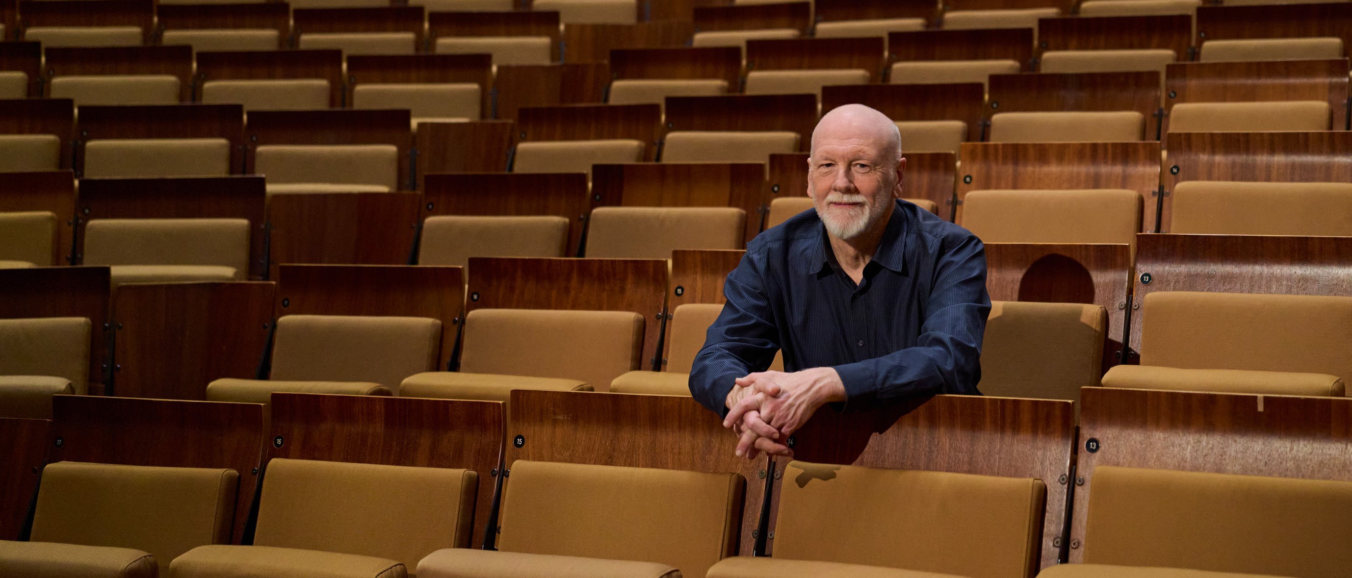 Brett Dean sitzt allein in der Mitte eines großen, leeren Saals mit braunen Sitzreihen und blickt nach vorne, die Arme auf den Sitz vor ihm gestützt.