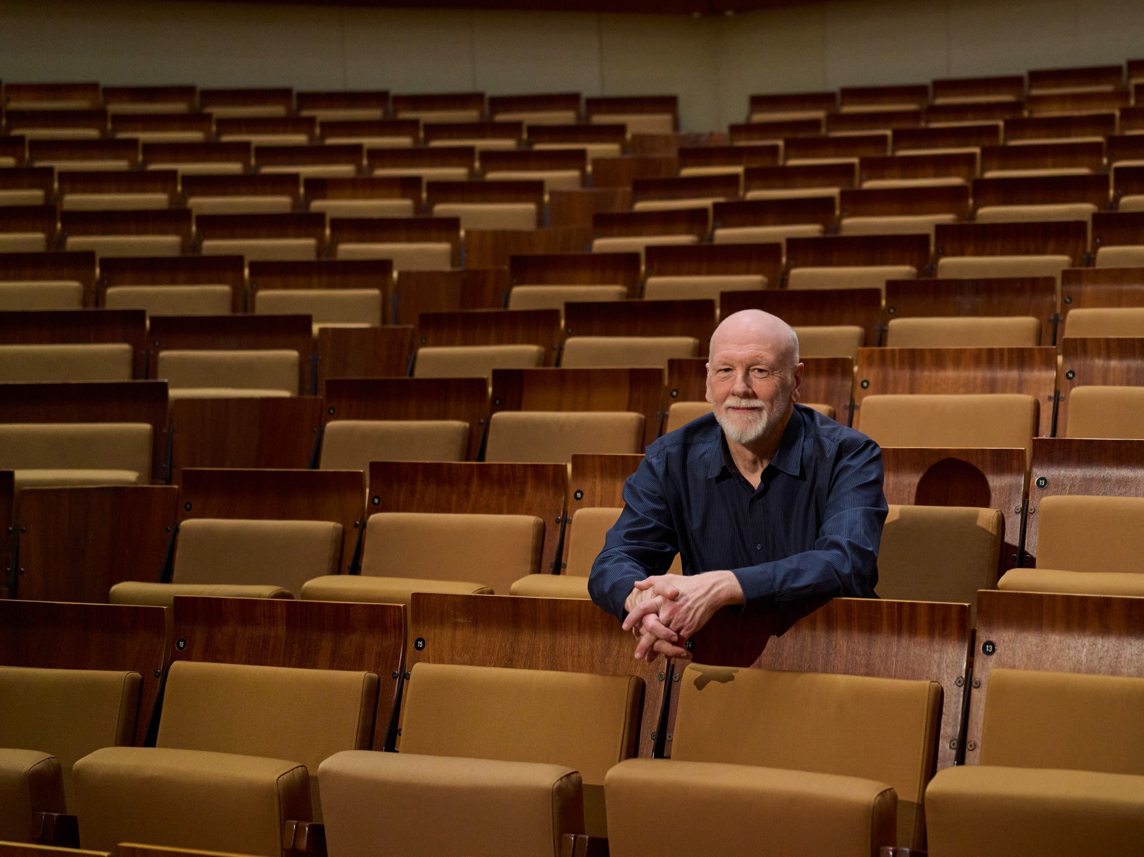 Brett Dean with a beard sits alone in the middle of many empty wooden auditorium seats, looking forward with his arms resting on the seat in front of him. The hall is empty and well-lit.