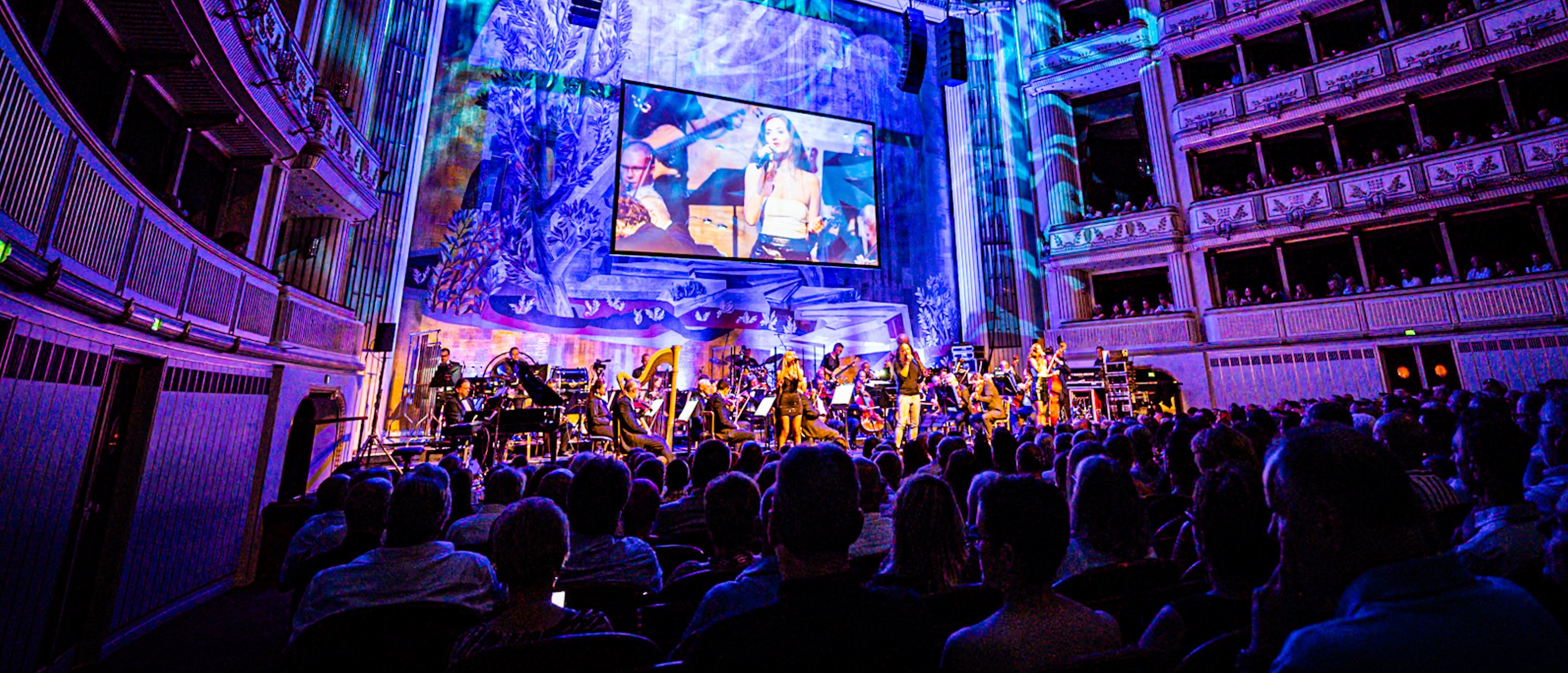 Eine lebhafte Konzertatmosphäre in einem Theater: Musiker treten auf der Bühne auf, bunte Lichter erhellen den Raum, und das Publikum sitzt in Reihen. Auf einer großen Leinwand ist eine Nahaufnahme eines Künstlers zu sehen.