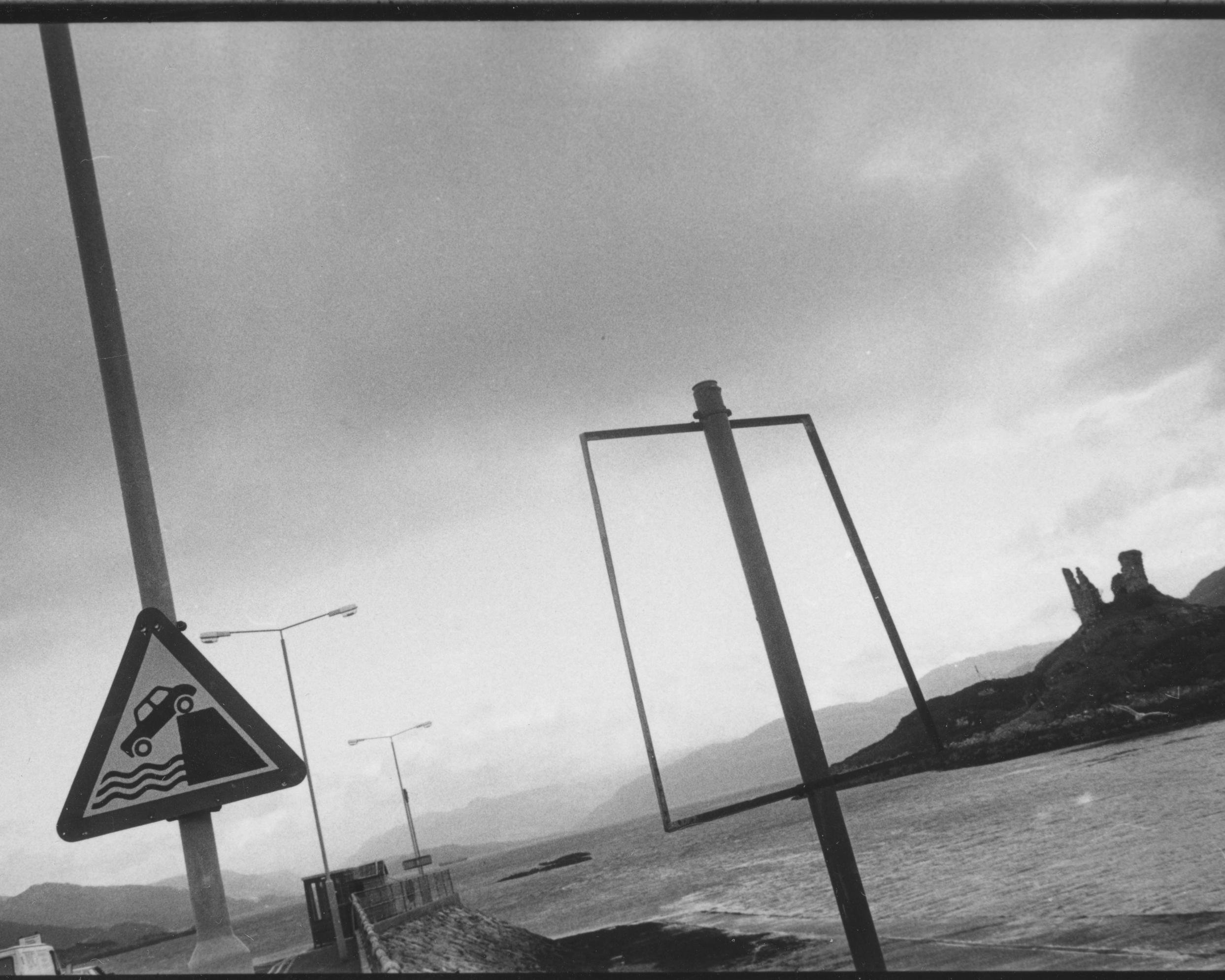 Ein Warnschild mit Küste, Meer und einem Auto, das ist Meer fällt, steht am Deich einer Bucht. Im Hintergrund Meer und weitere Landzungen.
