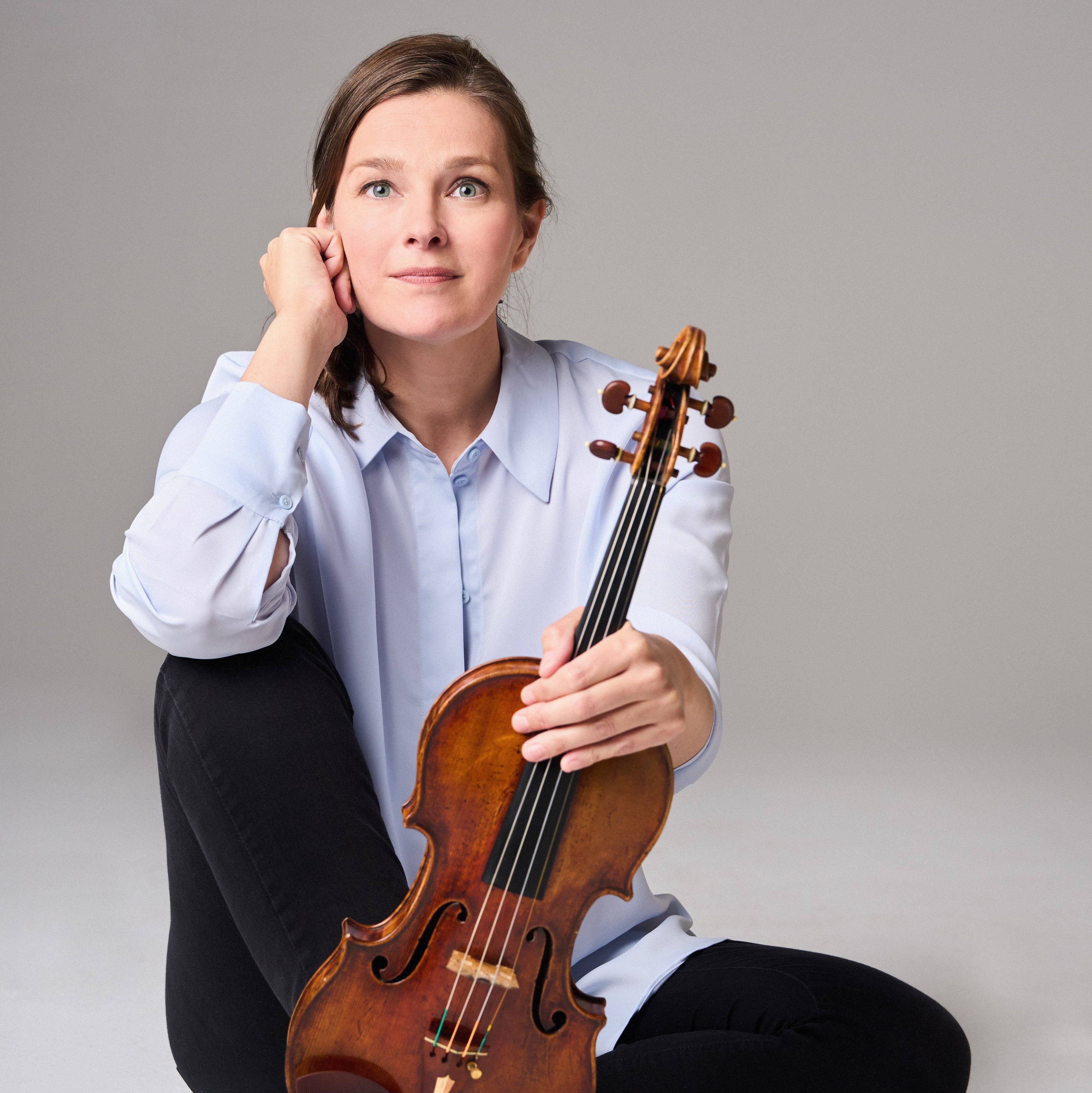 Janine Jansen with long hair, a light blue shirt and black trousers sits barefoot on the floor. She holds a violin upright on her foot and looks thoughtfully into the camera.
