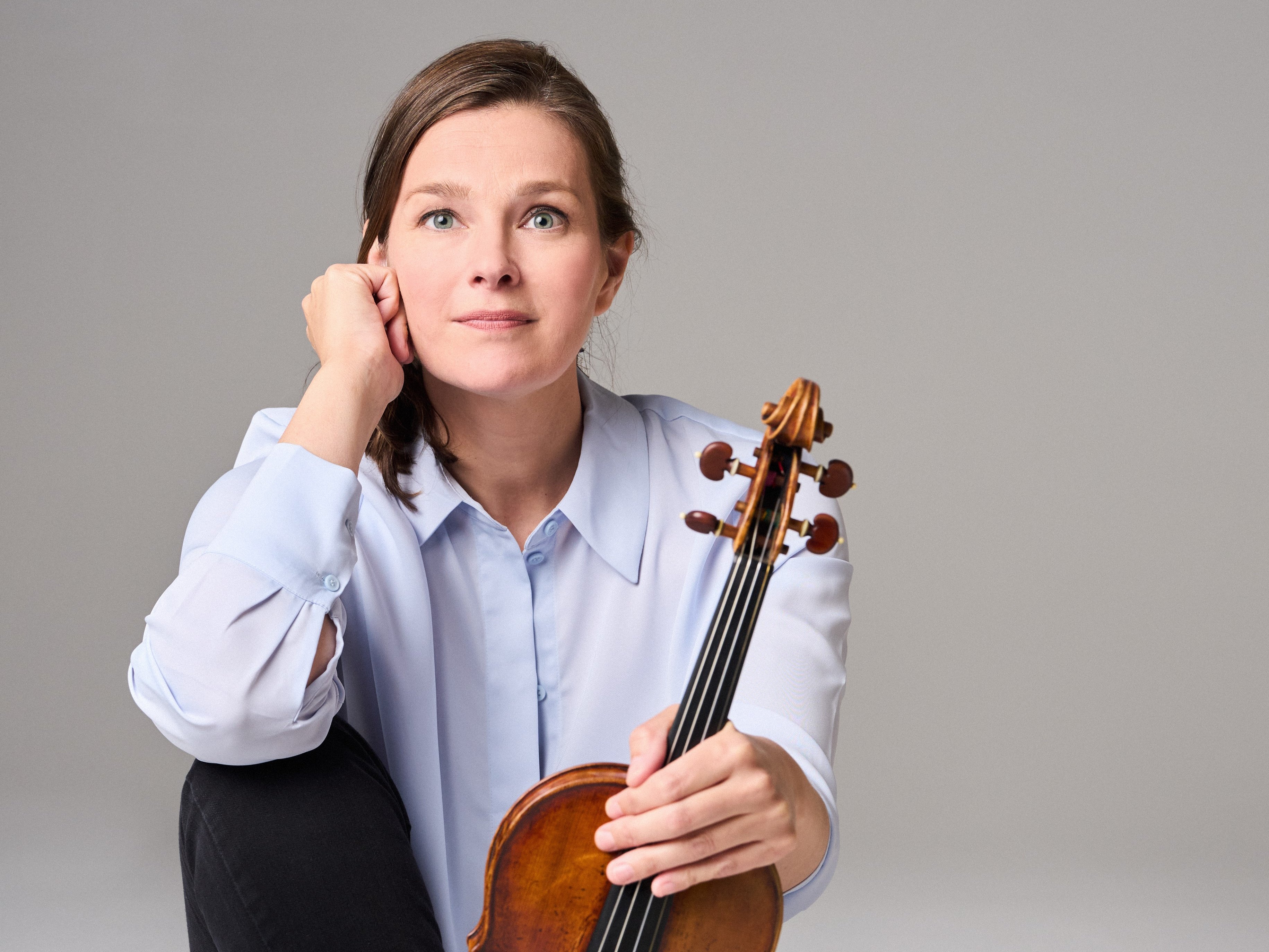 Janine Jansen mit langen Haaren, einem hellblauen Hemd und einer schwarzen Hose sitzt barfuß auf dem Boden. Sie hält eine Geige senkrecht auf ihrem Fuß und blickt nachdenklich in die Kamera.