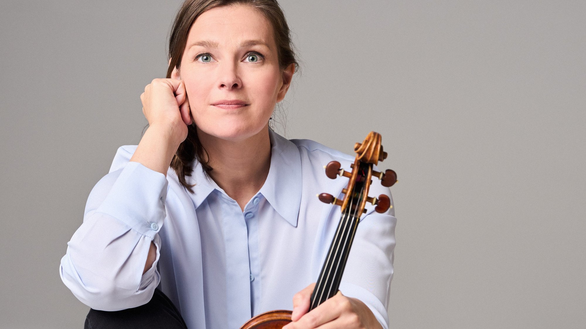Janine Jansen with long hair, a light blue shirt and black trousers sits barefoot on the floor. She holds a violin upright on her foot and looks thoughtfully into the camera.