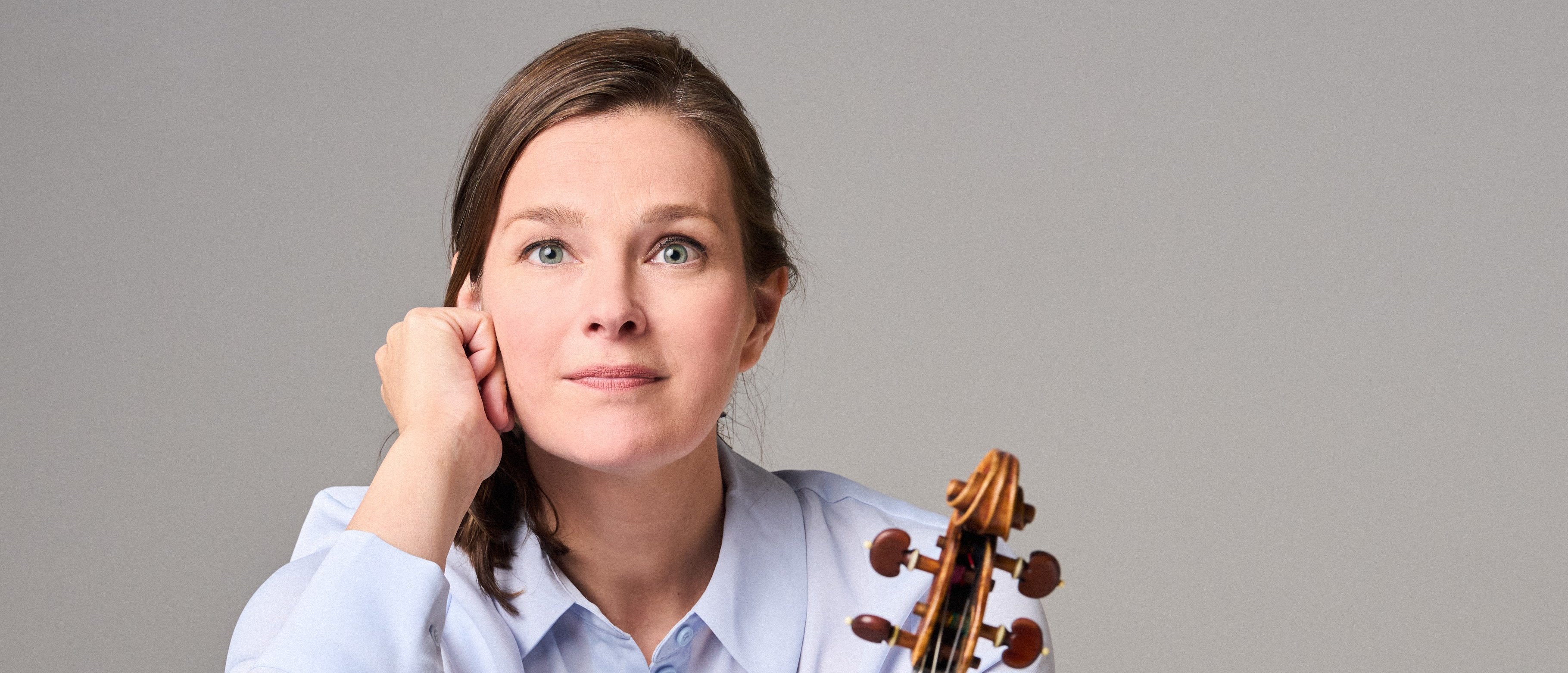 Janine Jansen mit langen Haaren, einem hellblauen Hemd und einer schwarzen Hose sitzt barfuß auf dem Boden. Sie hält eine Geige senkrecht auf ihrem Fuß und blickt nachdenklich in die Kamera.
