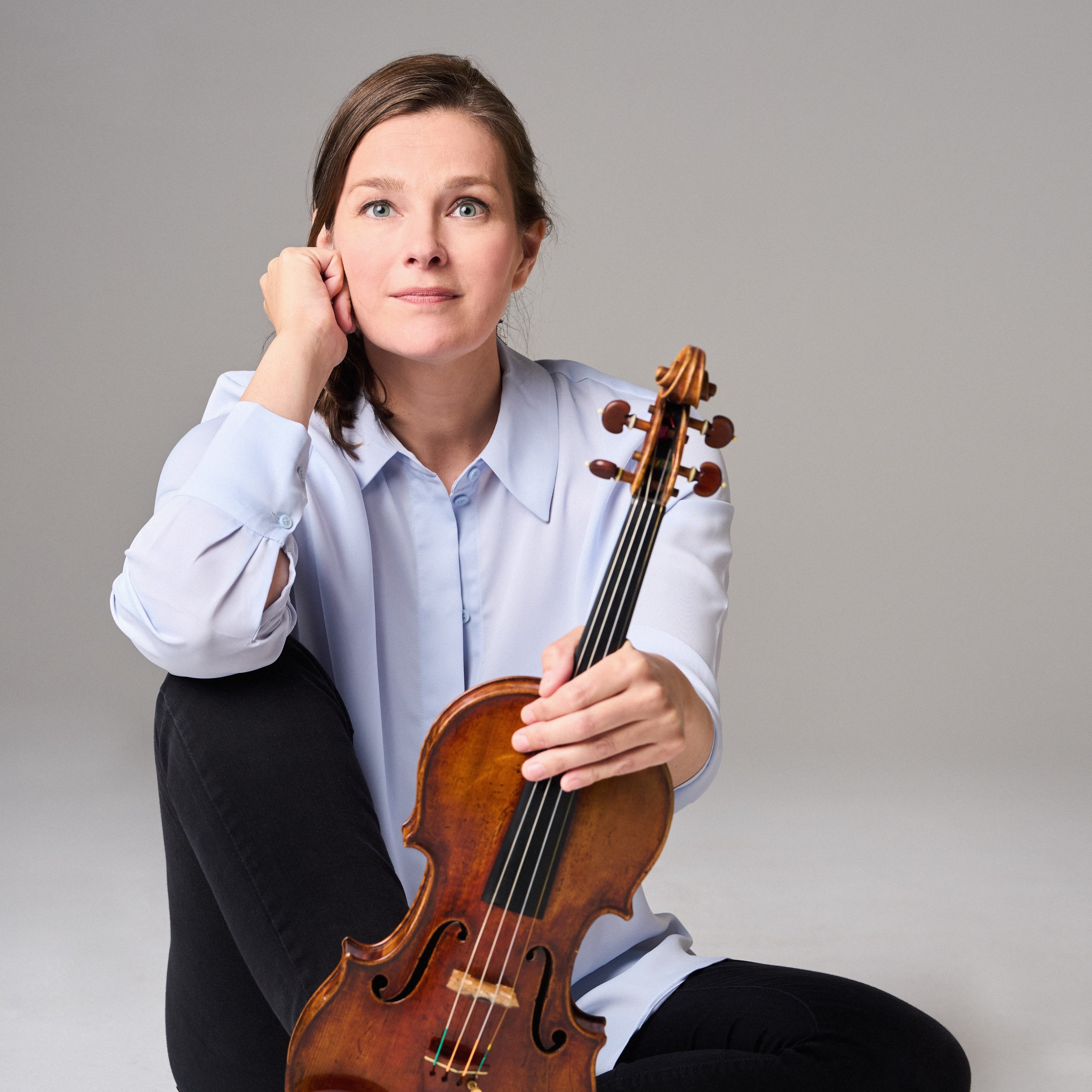 Janine Jansen with long hair, a light blue shirt and black trousers sits barefoot on the floor. She holds a violin upright on her foot and looks thoughtfully into the camera.