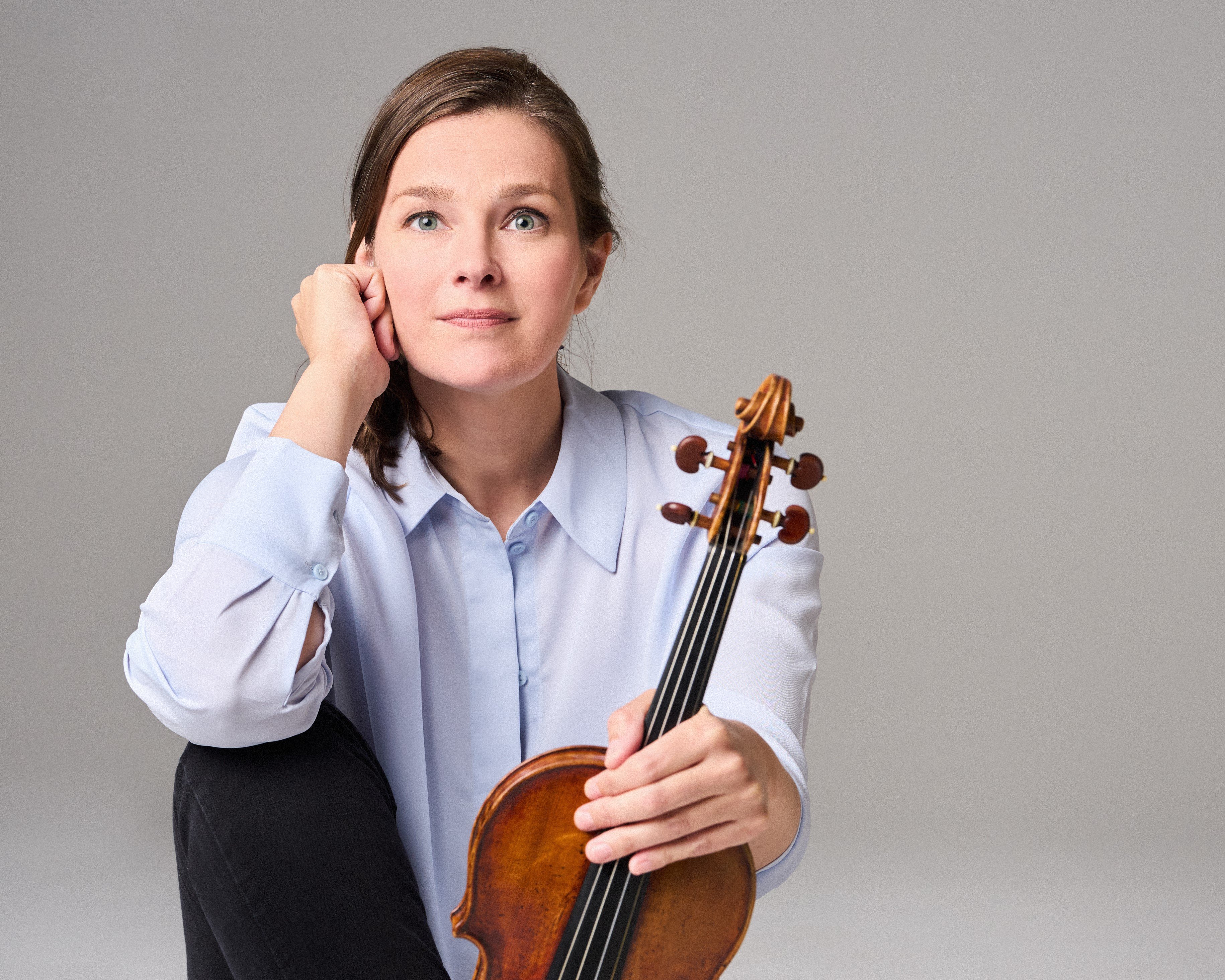 Janine Jansen mit langen Haaren, einem hellblauen Hemd und einer schwarzen Hose sitzt barfuß auf dem Boden. Sie hält eine Geige senkrecht auf ihrem Fuß und blickt nachdenklich in die Kamera.