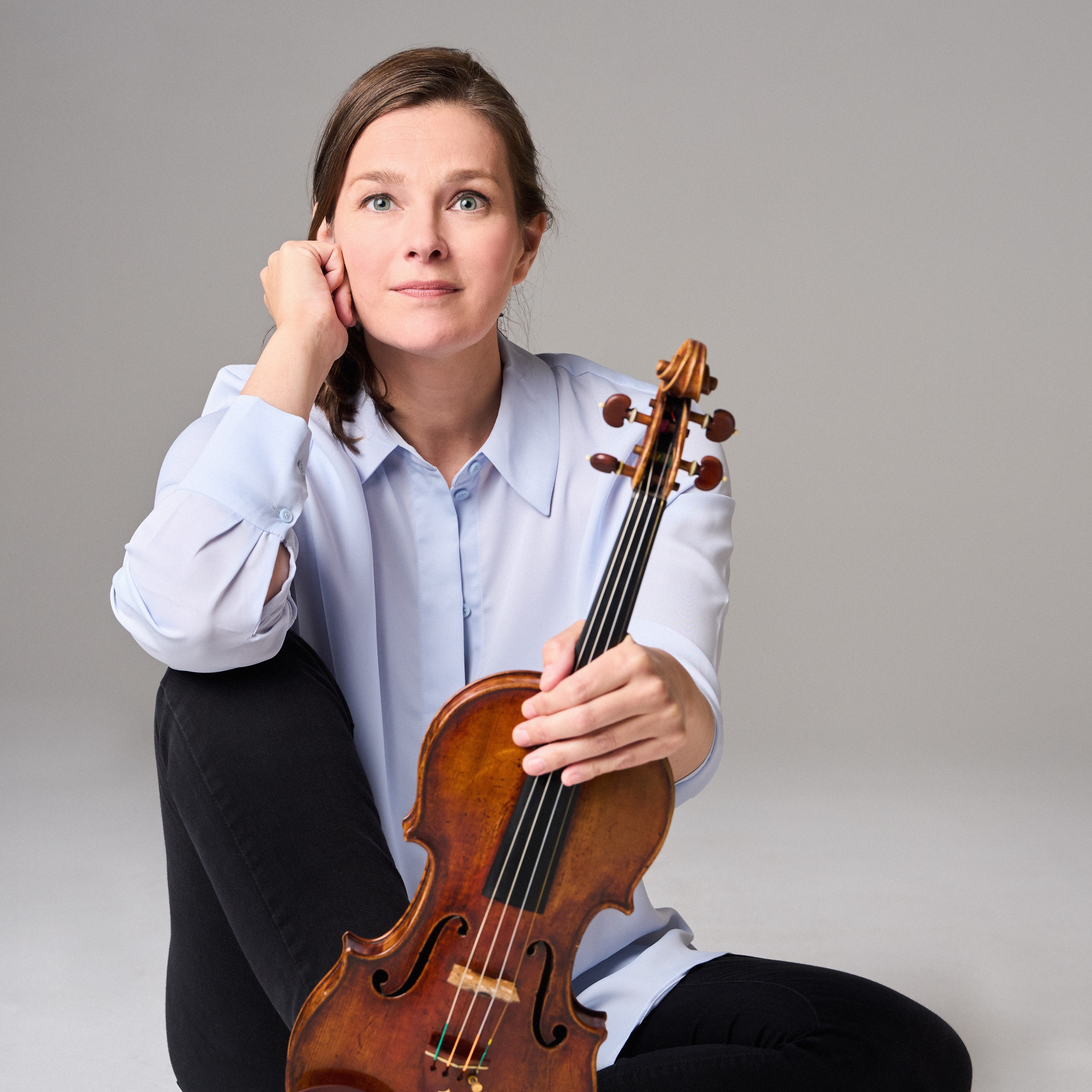 Janine Jansen mit langen Haaren, einem hellblauen Hemd und einer schwarzen Hose sitzt barfuß auf dem Boden. Sie hält eine Geige senkrecht auf ihrem Fuß und blickt nachdenklich in die Kamera.