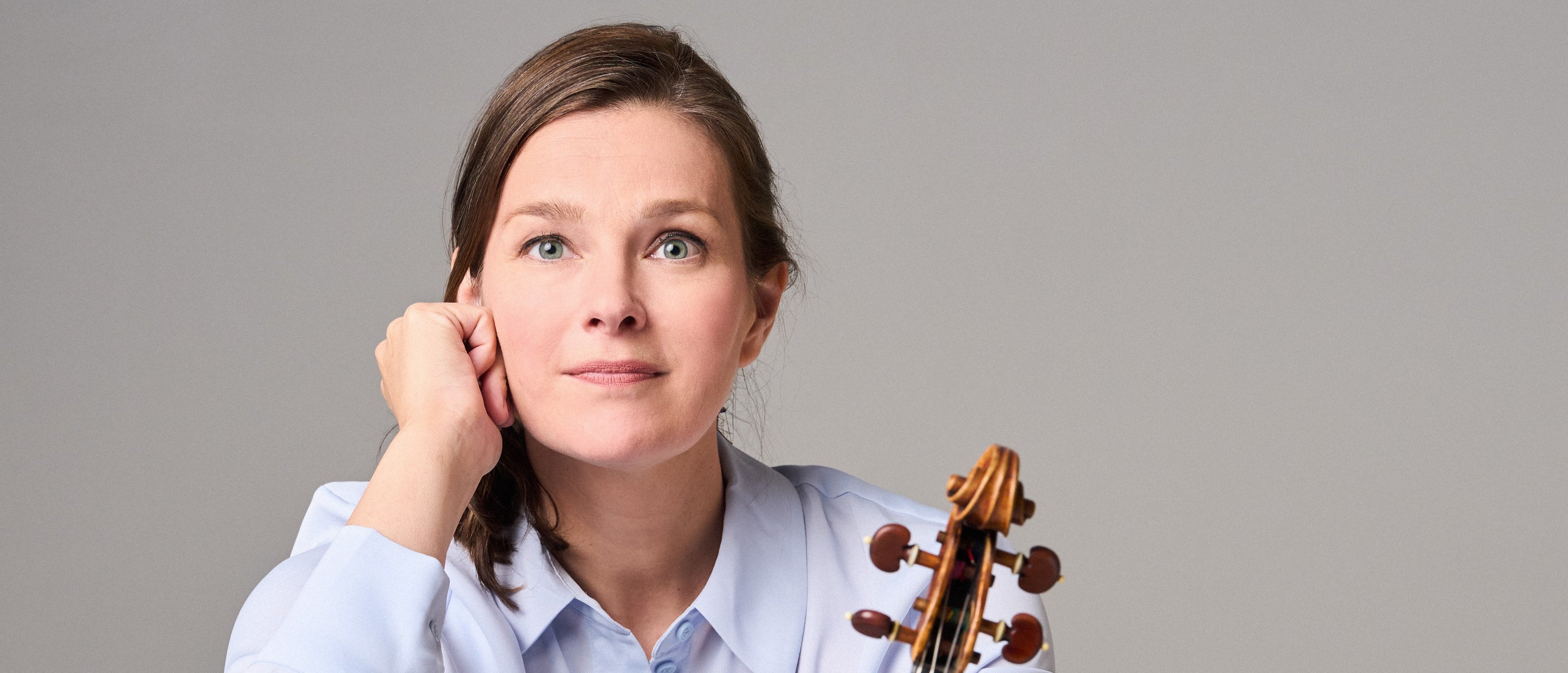 Janine Jansen mit langen Haaren, einem hellblauen Hemd und einer schwarzen Hose sitzt barfuß auf dem Boden. Sie hält eine Geige senkrecht auf ihrem Fuß und blickt nachdenklich in die Kamera.
