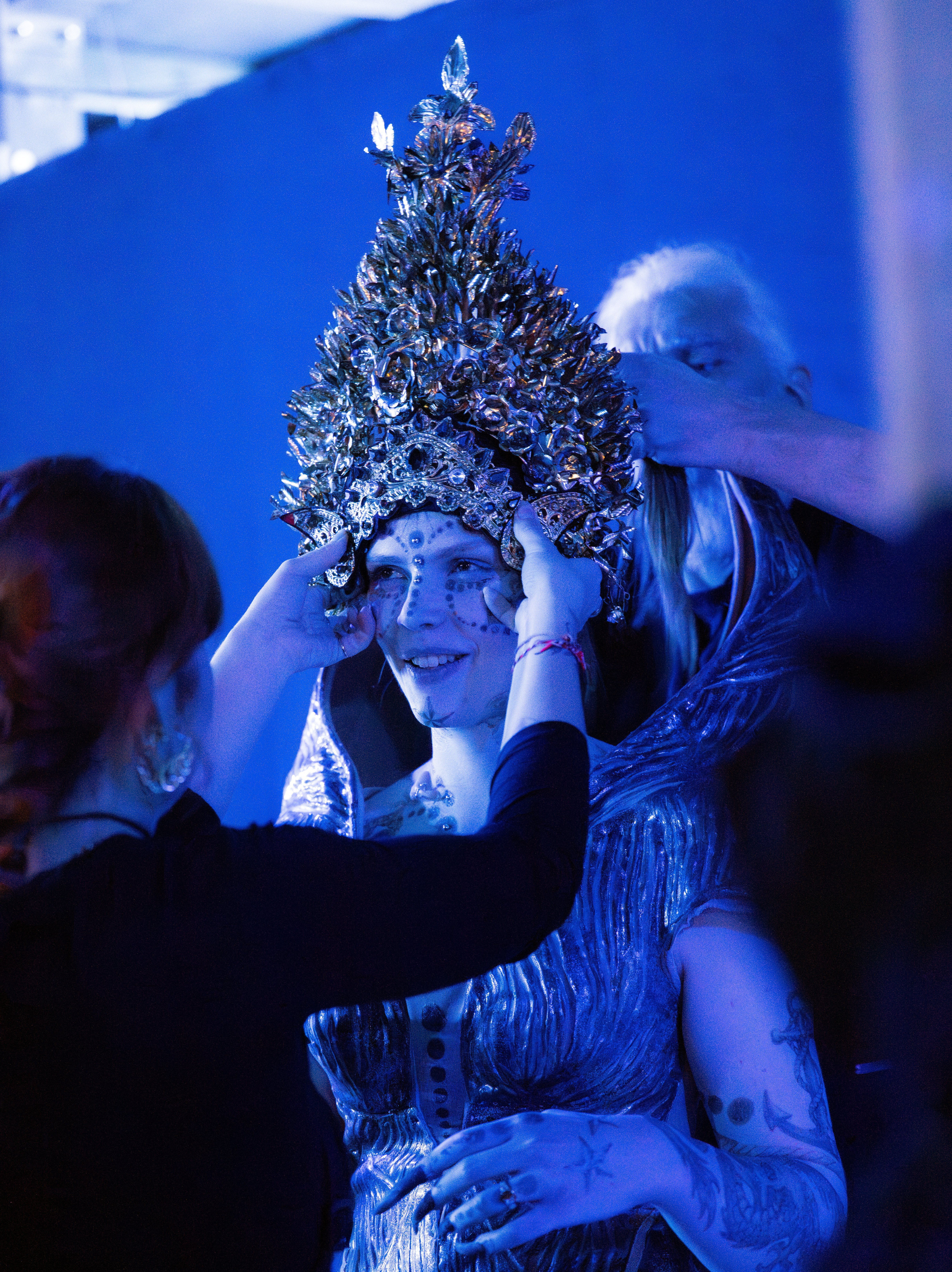 Eine Person mit einem verzierten, silbernen Kopfschmuck und einem schimmernden Outfit wird von zwei Personen unter blauem Licht unterstützt, die den Kopfschmuck und das Kostüm anpassen.