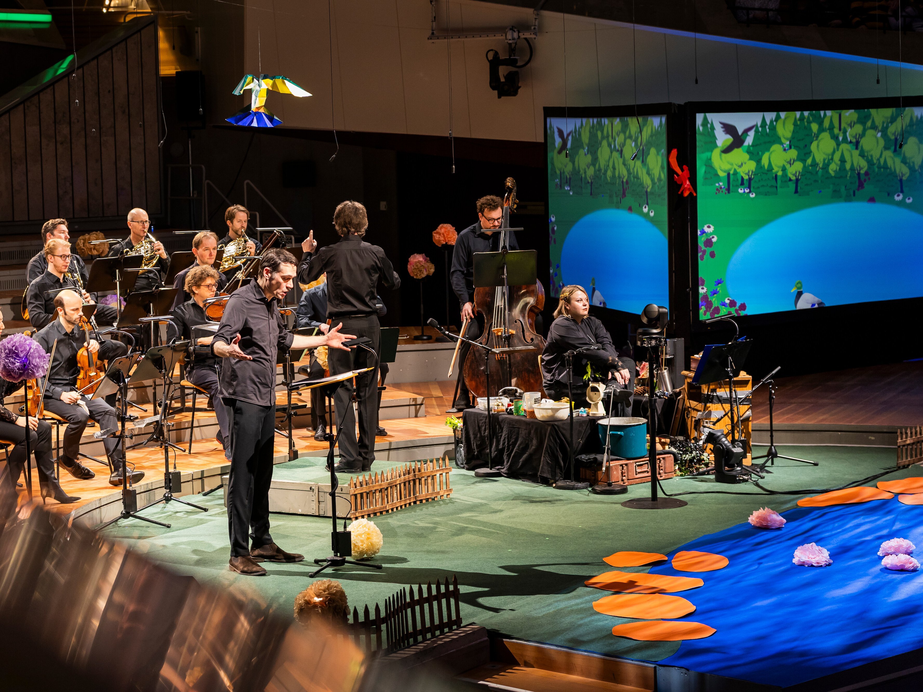 Ein Mann singt mit Musikern auf der Bühne, während hinter ihnen animierte Naturszenen projiziert werden; die Bühne ist mit künstlichen Blumen und einem Teich geschmückt.