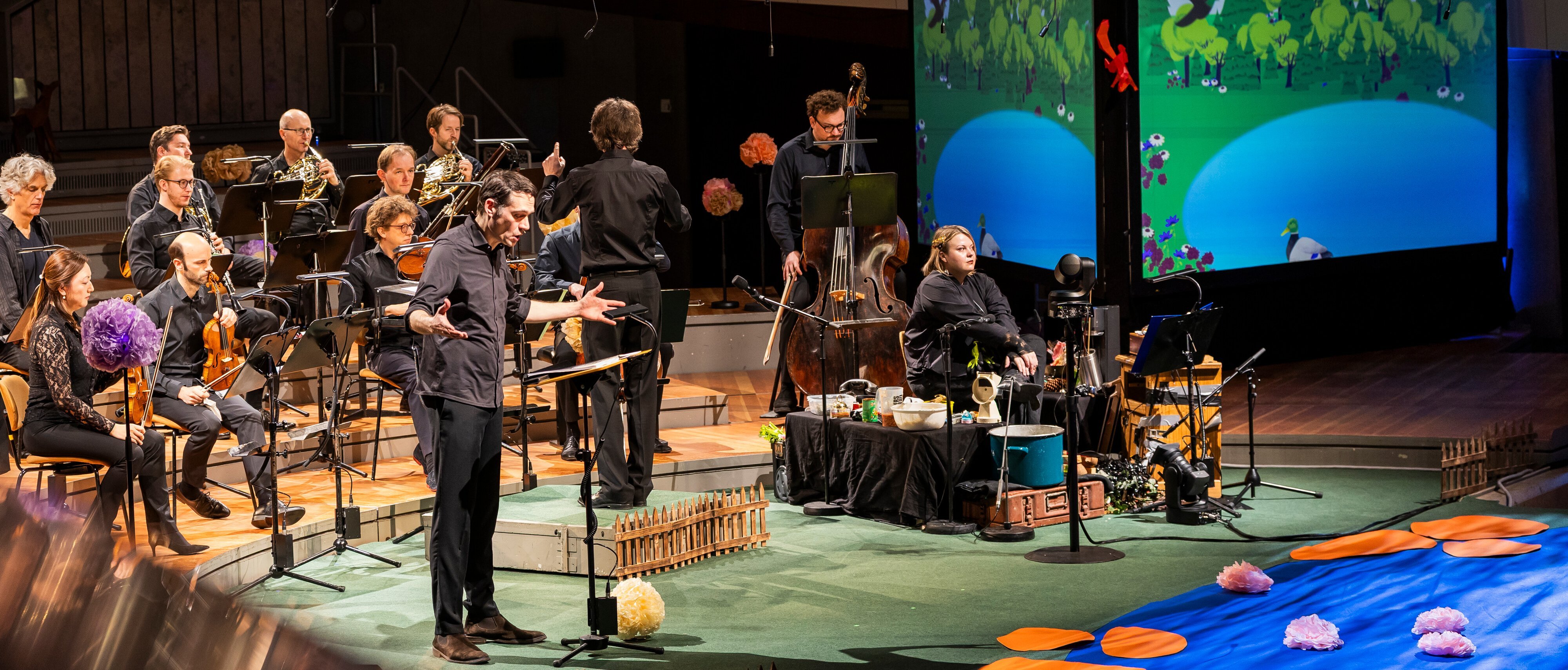 Ein Mann singt mit Musikern auf der Bühne, während hinter ihnen animierte Naturszenen projiziert werden; die Bühne ist mit künstlichen Blumen und einem Teich geschmückt.
