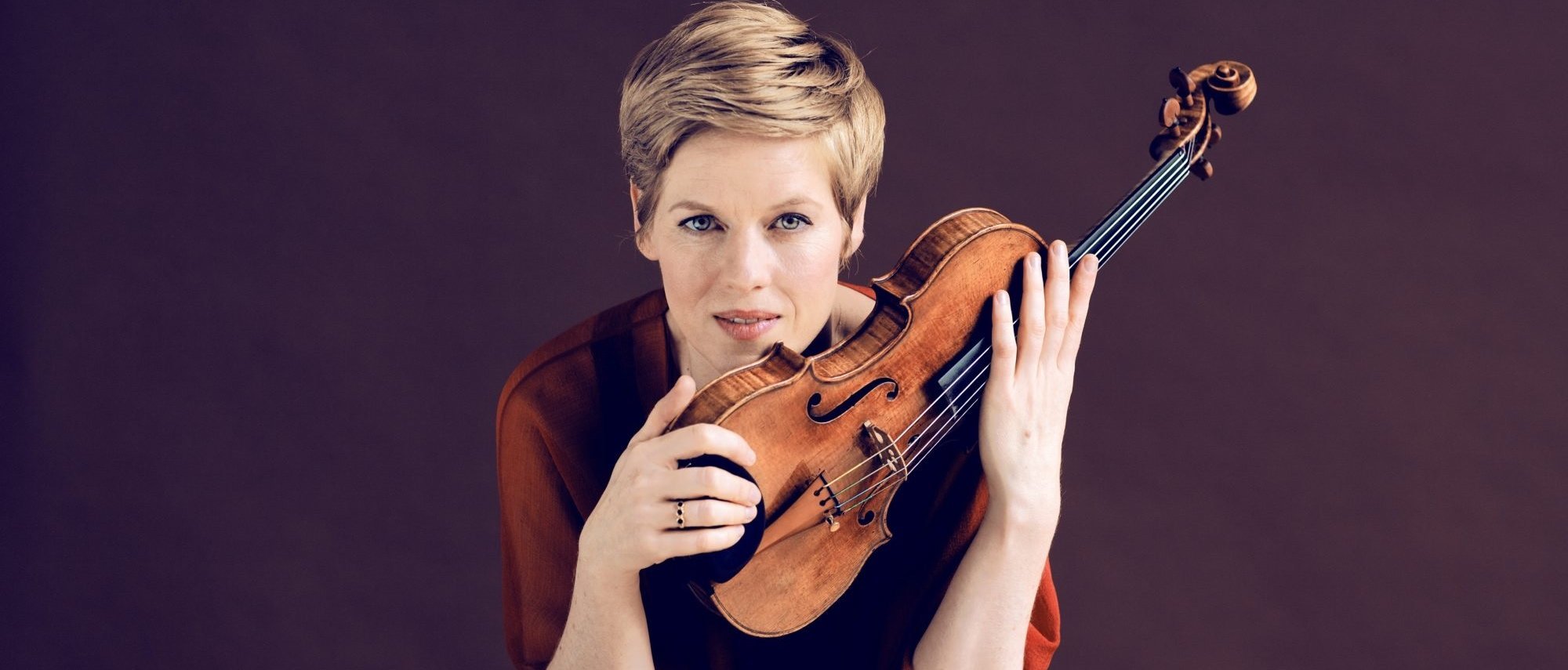 Isabelle Faust mit kurzen blonden Haaren und ernster Miene hält vor einem schlichten dunklen Hintergrund eine Geige aufrecht vor ihr Gesicht.