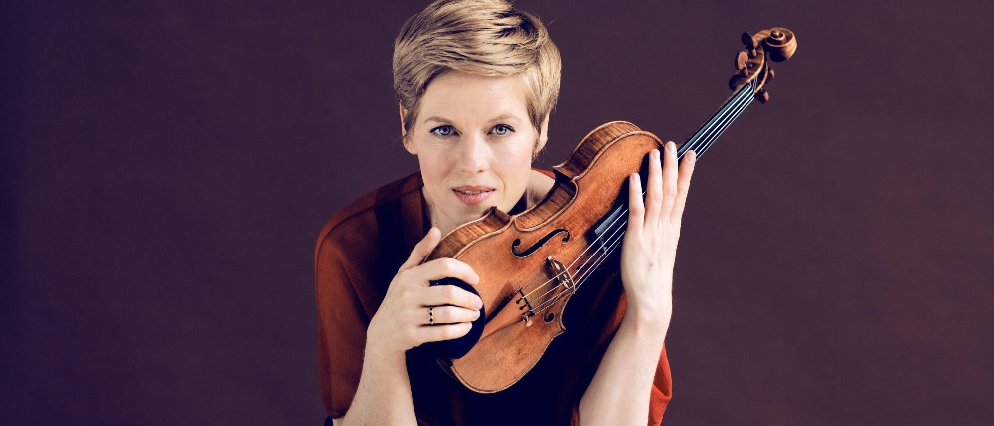 Isabelle Faust mit kurzen blonden Haaren und ernster Miene hält vor einem schlichten dunklen Hintergrund eine Geige aufrecht vor ihr Gesicht.