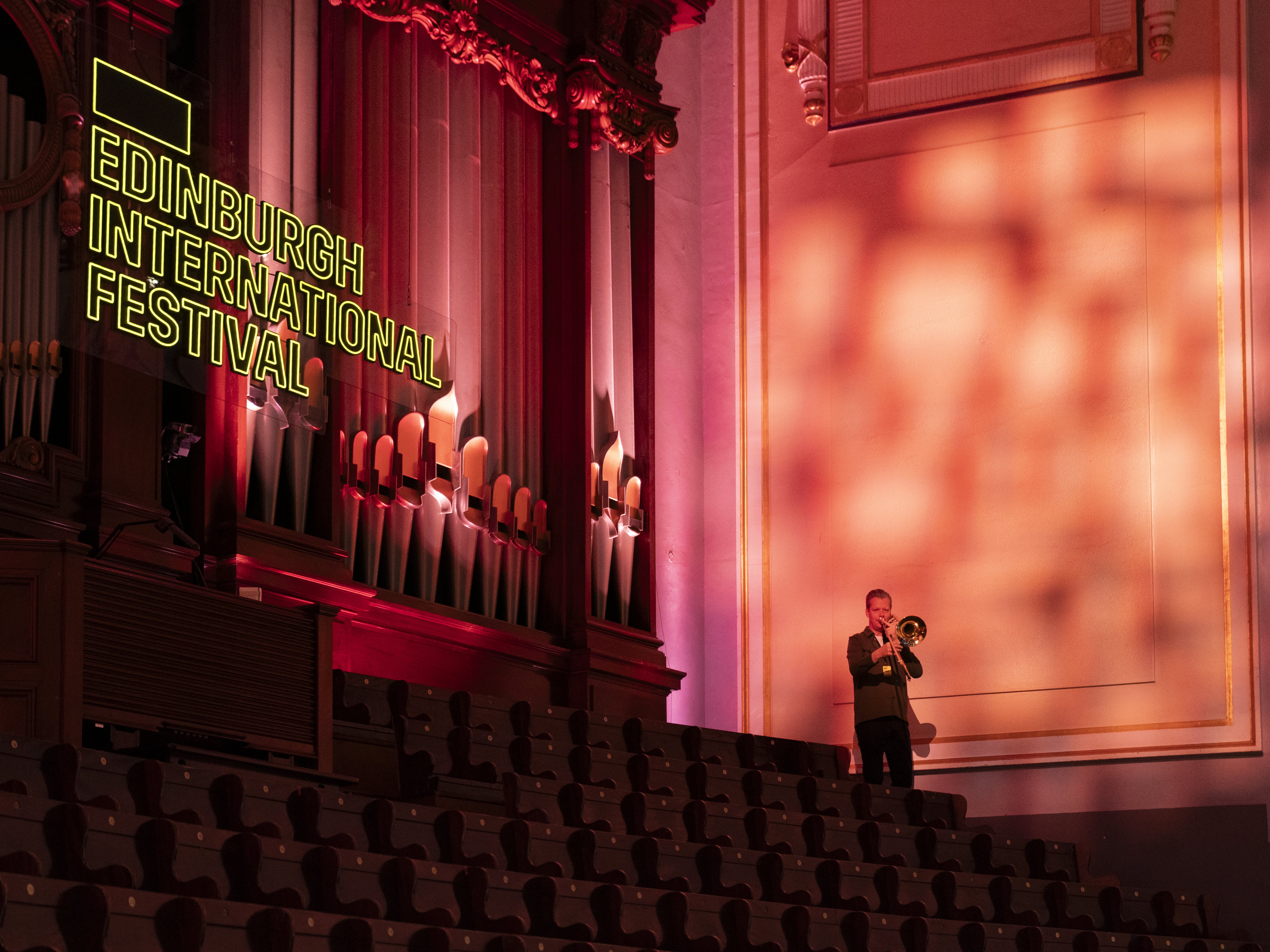 A musician plays a trombone in an empty, ornate hall with a pipe organ. Neon Edinburgh International Festival sign glows on the wall. Warm lighting creates dramatic shadows on the right.