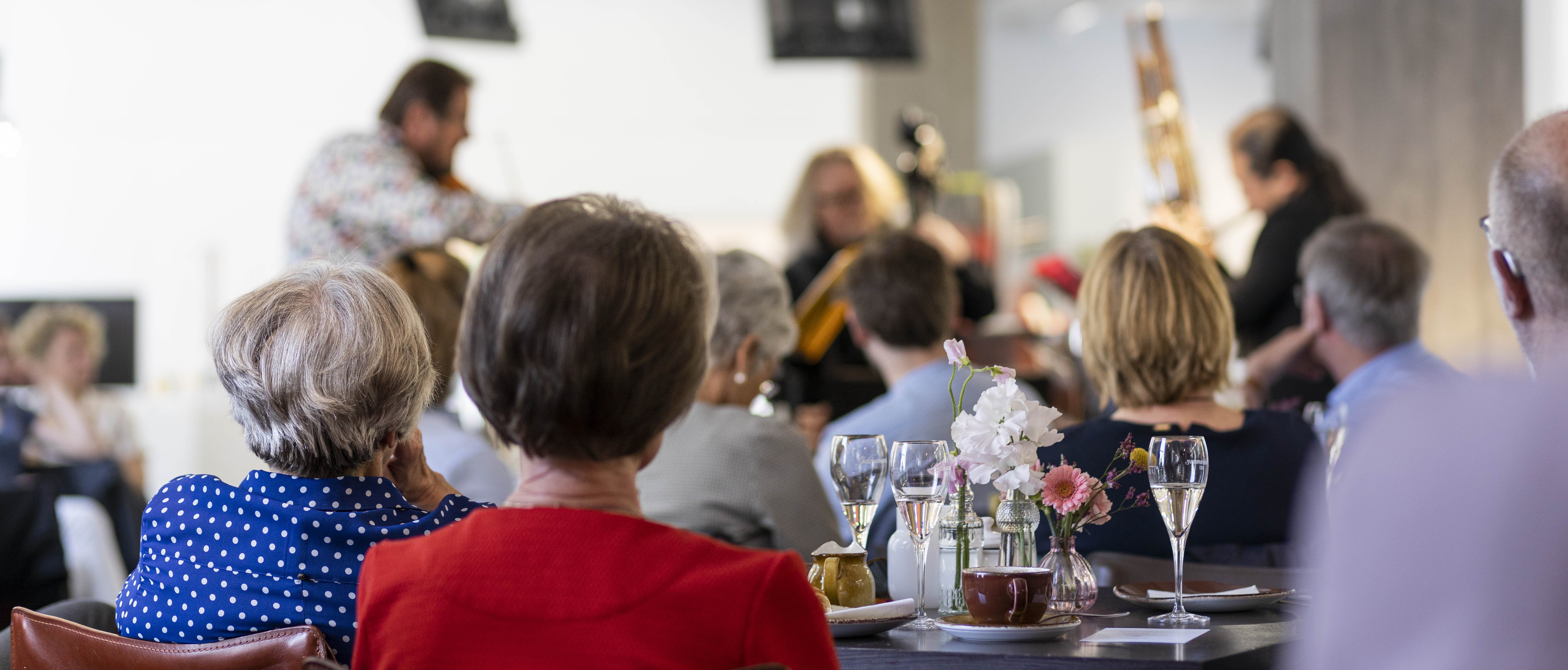 Menschen, die an einem Tisch mit Getränken und Blumen sitzen, sehen Musikern zu, die in einem hellen, modernen Raum auftreten. Der Fokus liegt auf dem Publikum, während die Musiker im Hintergrund verschwimmen.