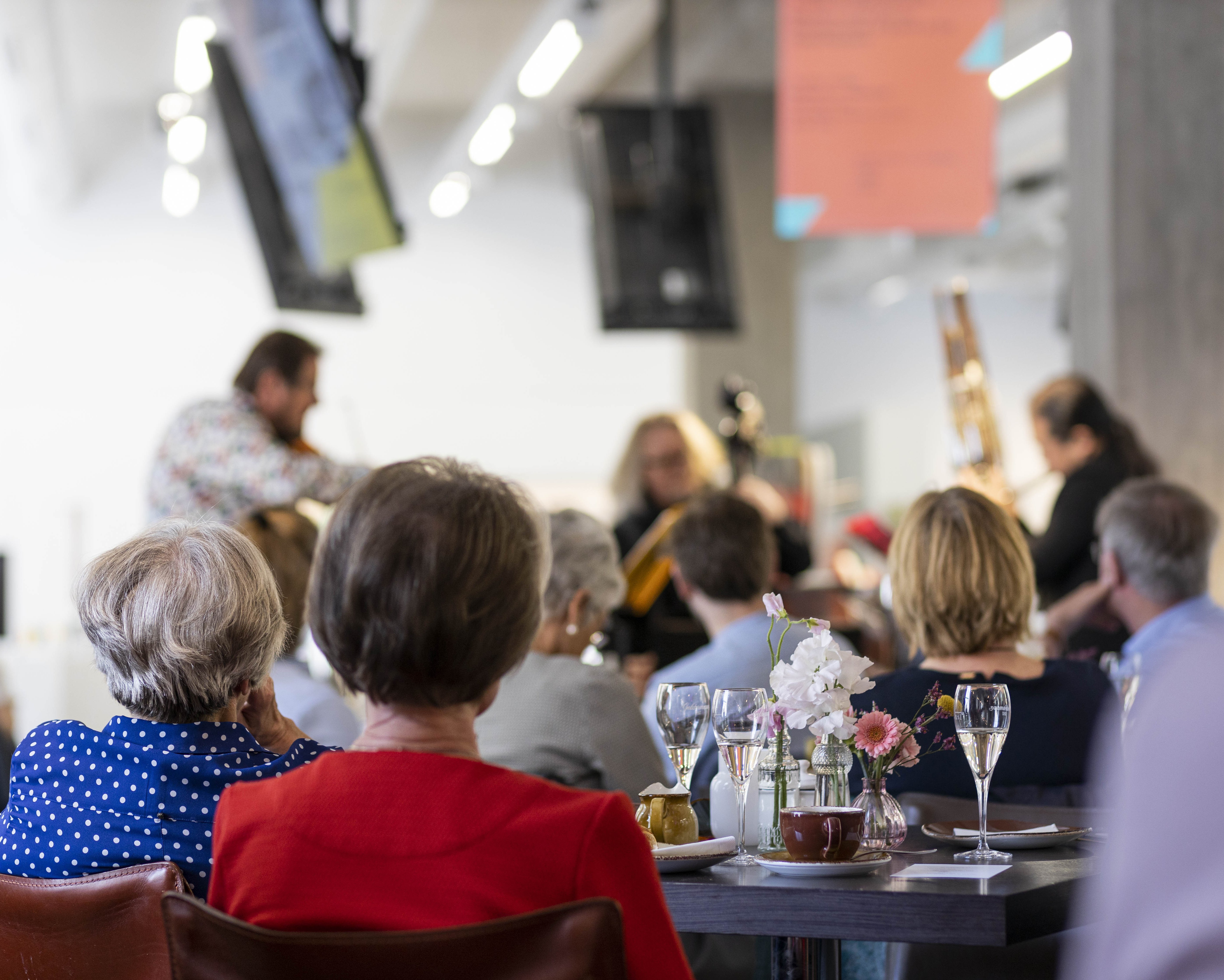 Menschen, die an einem Tisch mit Getränken und Blumen sitzen, sehen Musikern zu, die in einem hellen, modernen Raum auftreten. Der Fokus liegt auf dem Publikum, während die Musiker im Hintergrund verschwimmen.