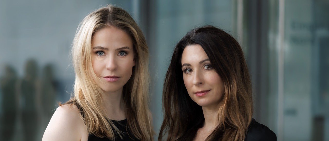 Two women with long hair, one blonde and one brunette, stand side by side wearing black tops, looking directly at the camera with a neutral expression. The background is blurred and modern.
