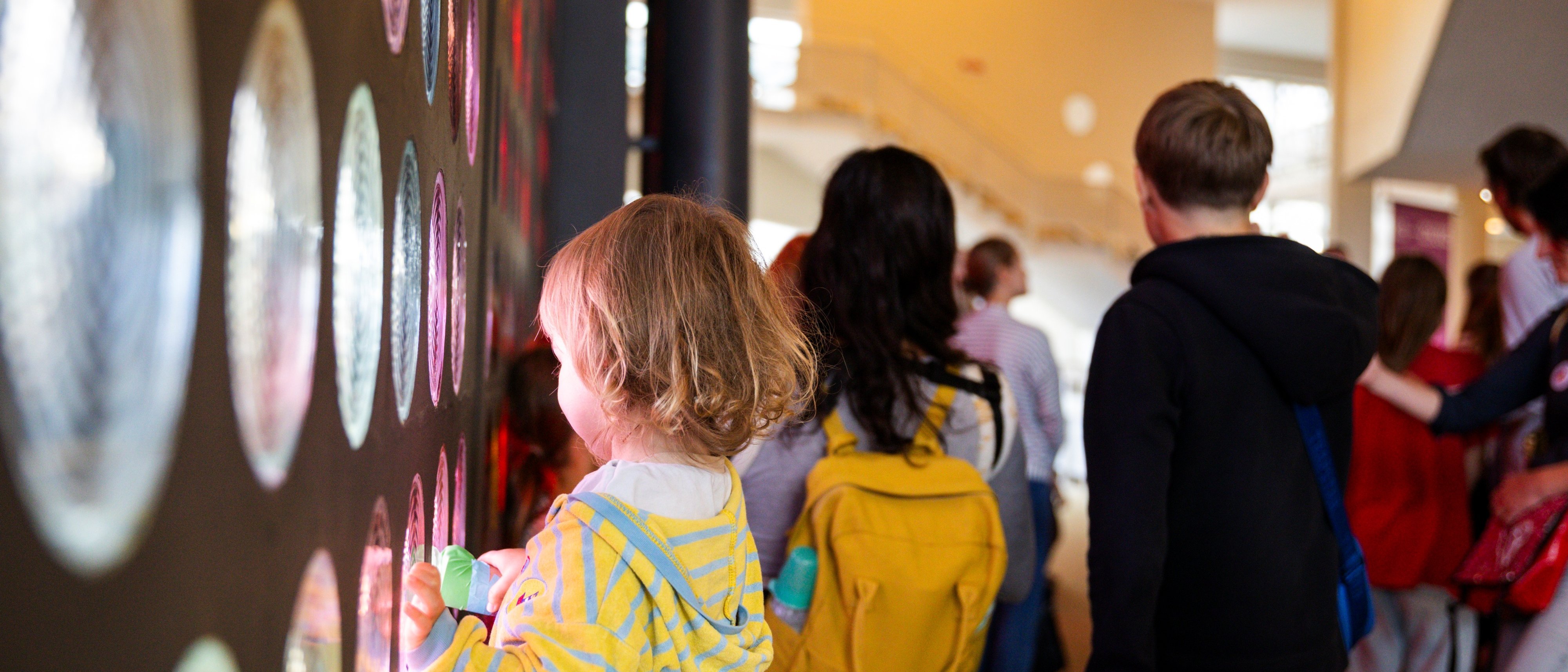 Ein Kind interagiert mit einer Wand aus bunten, kreisförmigen Glasbausteinen in einem hellen Innenraum. Das Kind im Vordergrund trägt einen gelb gestreiften Kapuzenpullover und berührt das Glas, während andere Personen in der Nähe stehen.