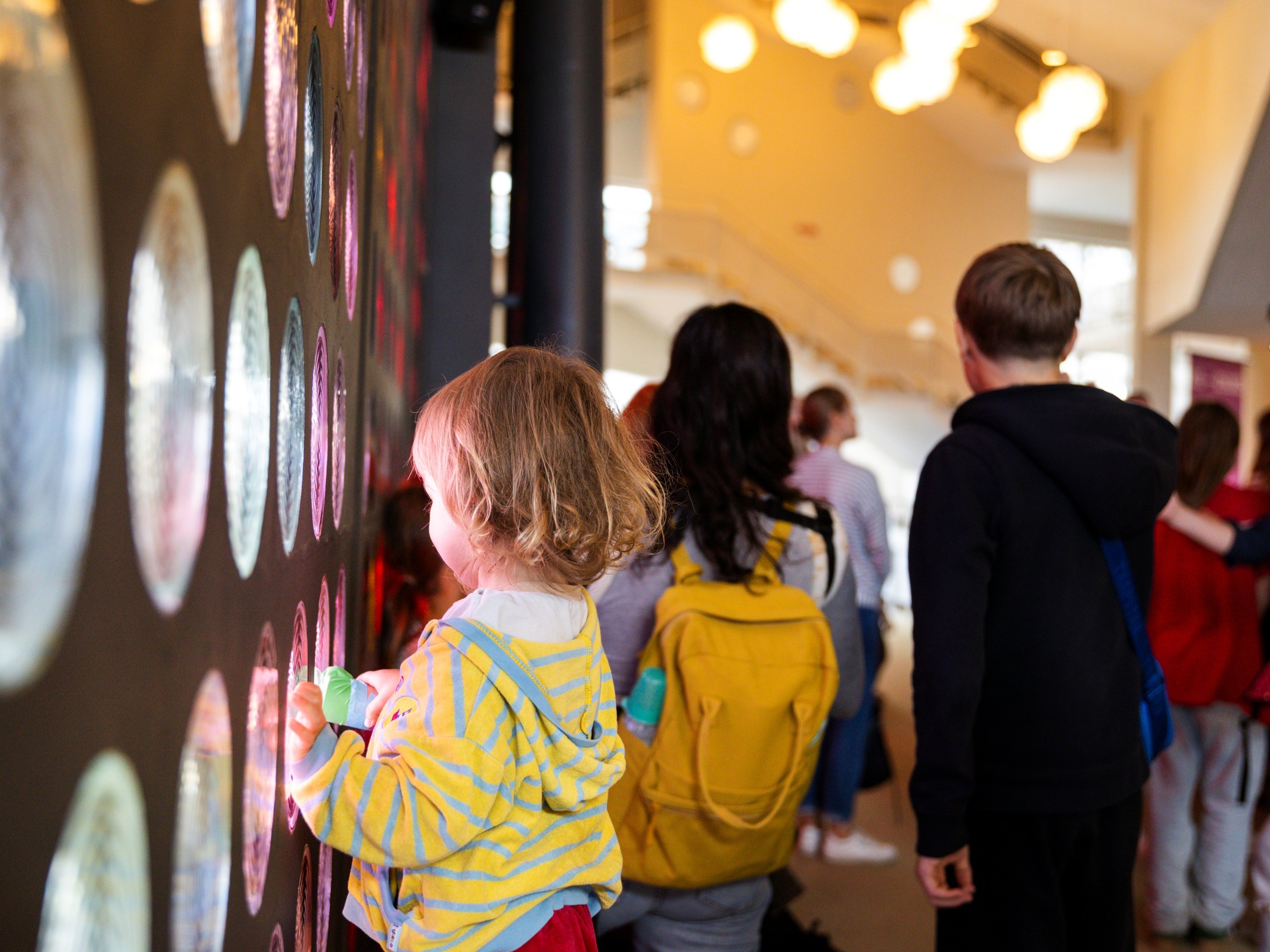 Ein Kind interagiert mit einer Wand aus bunten, kreisförmigen Glasbausteinen in einem hellen Innenraum. Das Kind im Vordergrund trägt einen gelb gestreiften Kapuzenpullover und berührt das Glas, während andere Personen in der Nähe stehen.