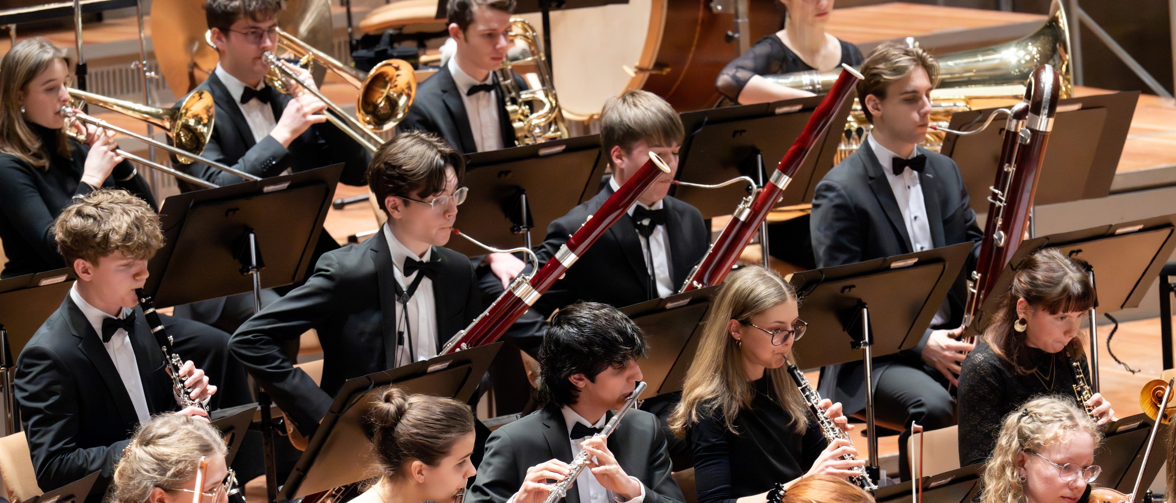 Ein junges Sinfonieorchester spielt auf der Bühne. Die Musiker spielen Holzblas- und Streichinstrumente und sind alle in formelle schwarze Kleidung gekleidet. Die Notenblätter liegen auf Ständern vor ihnen.