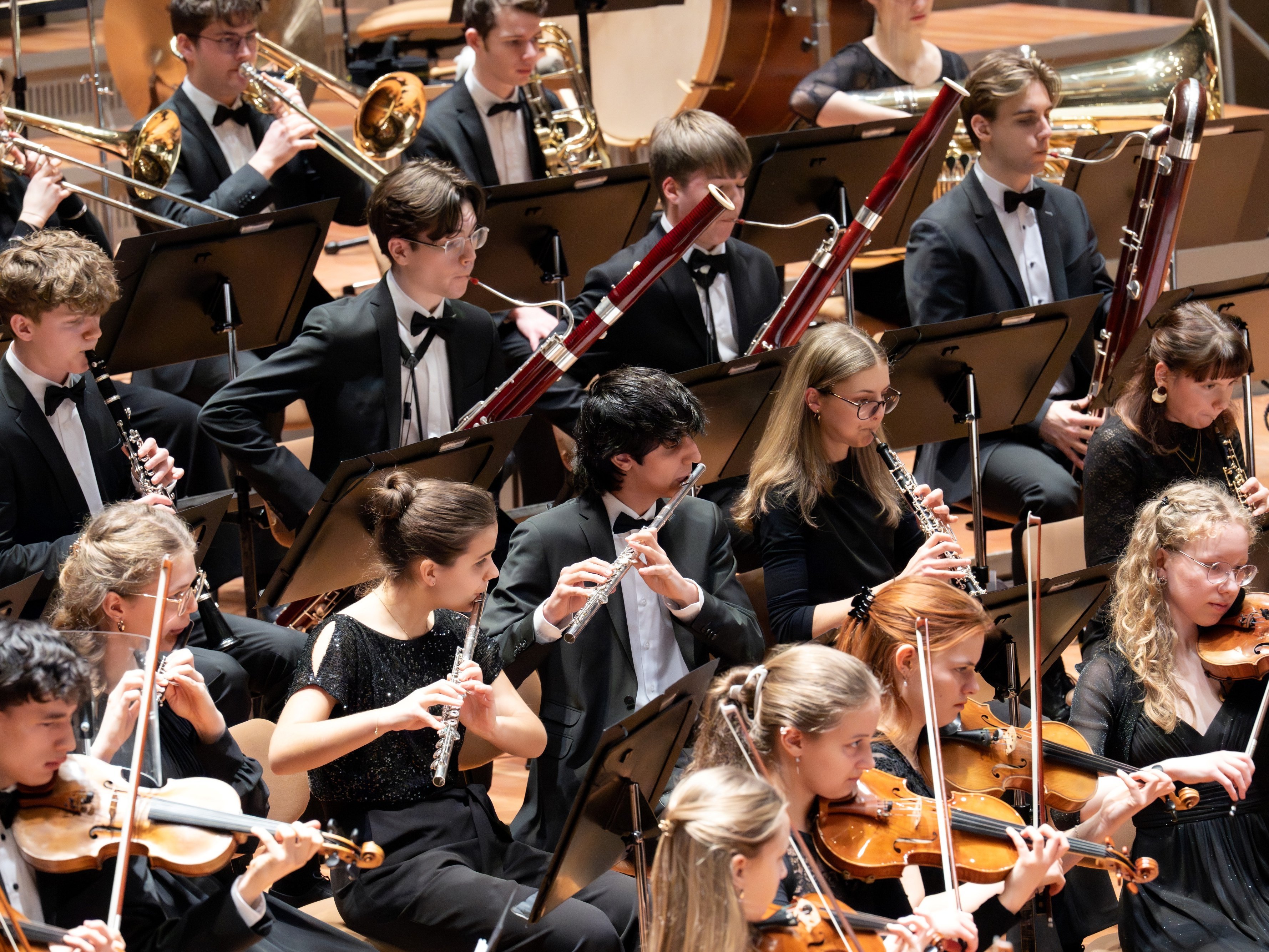 Ein junges Sinfonieorchester spielt auf der Bühne. Die Musiker spielen Holzblas- und Streichinstrumente und sind alle in formelle schwarze Kleidung gekleidet. Die Notenblätter liegen auf Ständern vor ihnen.