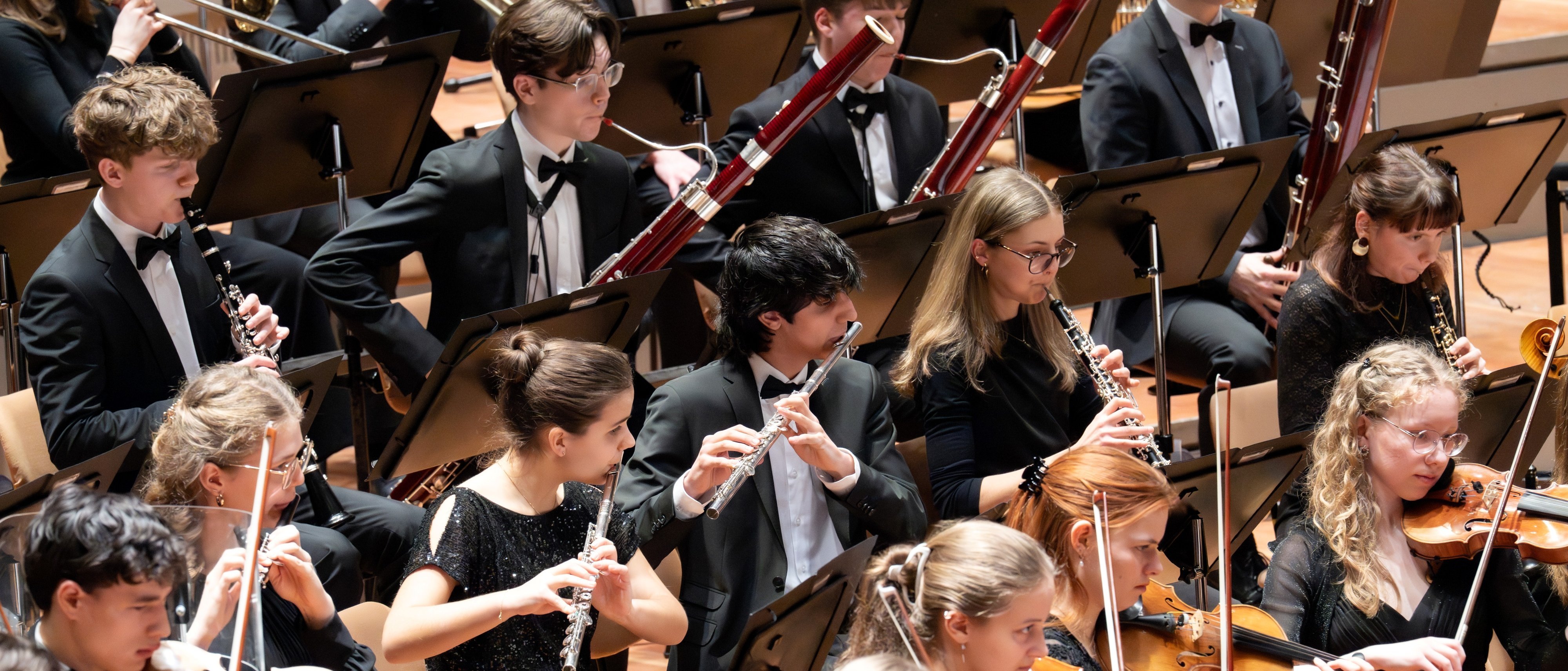 Ein junges Sinfonieorchester spielt auf der Bühne. Die Musiker spielen Holzblas- und Streichinstrumente und sind alle in formelle schwarze Kleidung gekleidet. Die Notenblätter liegen auf Ständern vor ihnen.