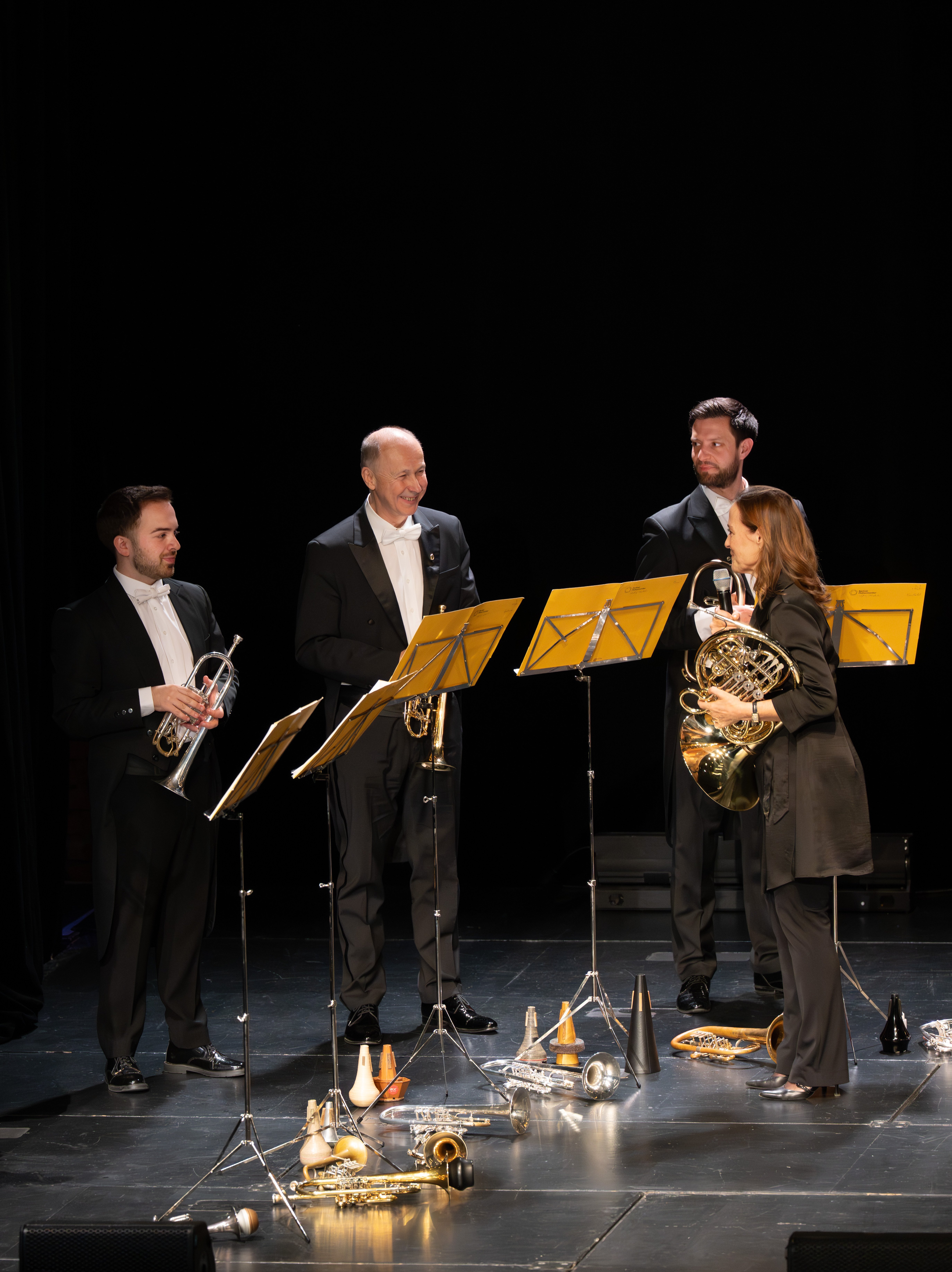 Vier Musiker in förmlicher Kleidung stehen mit Blasinstrumenten und Notenständern auf der Bühne, unterhalten sich und lächeln. Um sie herum sind verschiedene Blechblasinstrumente und Dämpfer auf dem Boden verteilt.