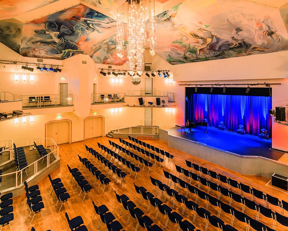 Ein helles Theater mit Reihen blauer Stühle, die auf eine Bühne mit blauen Vorhängen ausgerichtet sind, einem großen Kronleuchter, einer bunt bemalten Decke und Sitzplätzen auf beiden Seiten des Balkons.