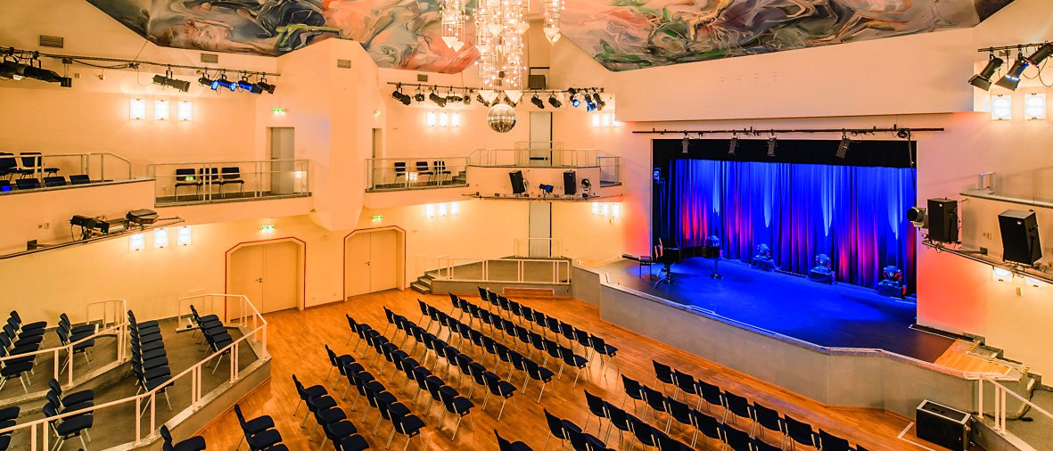 Ein helles Theater mit Reihen blauer Stühle, die auf eine Bühne mit blauen Vorhängen ausgerichtet sind, einem großen Kronleuchter, einer bunt bemalten Decke und Sitzplätzen auf beiden Seiten des Balkons.