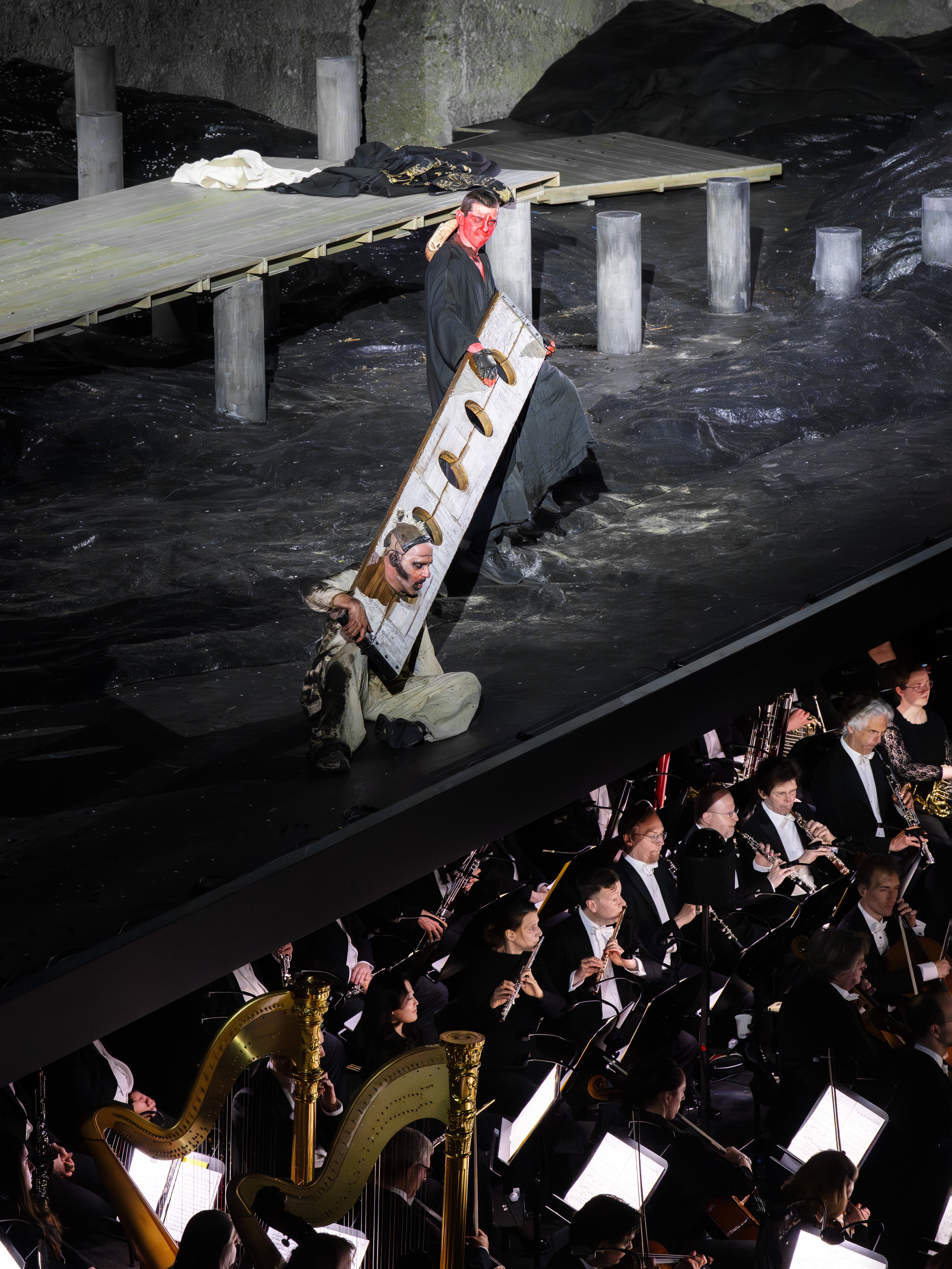 Ein Bühnenkünstler mit Maske steht auf einem schwarzen Bühnenbild mit einem Holzsteg. Unten spielt ein Orchester mit Harfen und Musikern Musik.