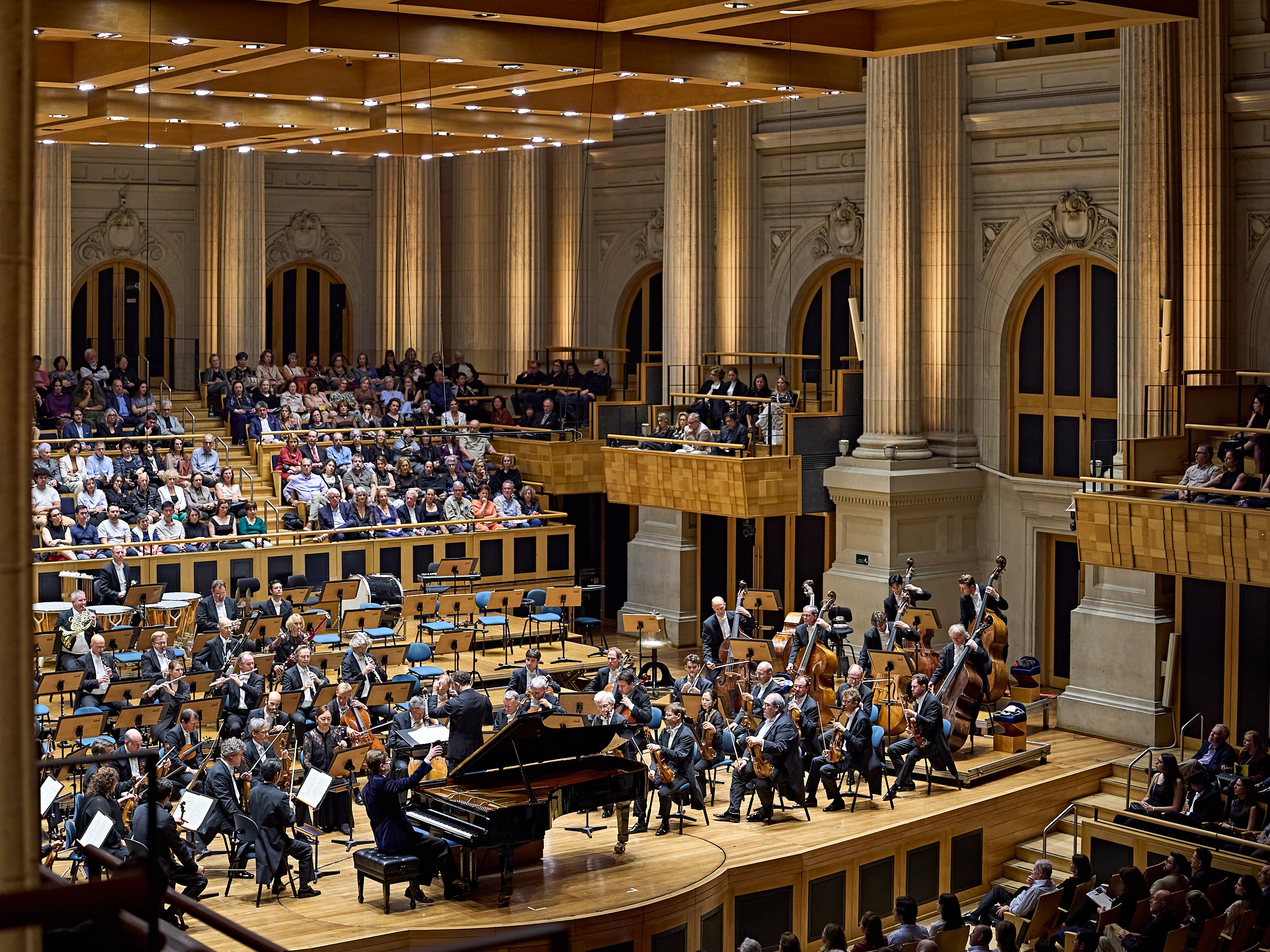 Ein Orchester spielt auf der Bühne eines prächtigen Konzertsaals, im Vordergrund sitzt ein Pianist am Flügel, und hinter ihm sowie an den Seiten sitzt ein großes Publikum in Reihen.