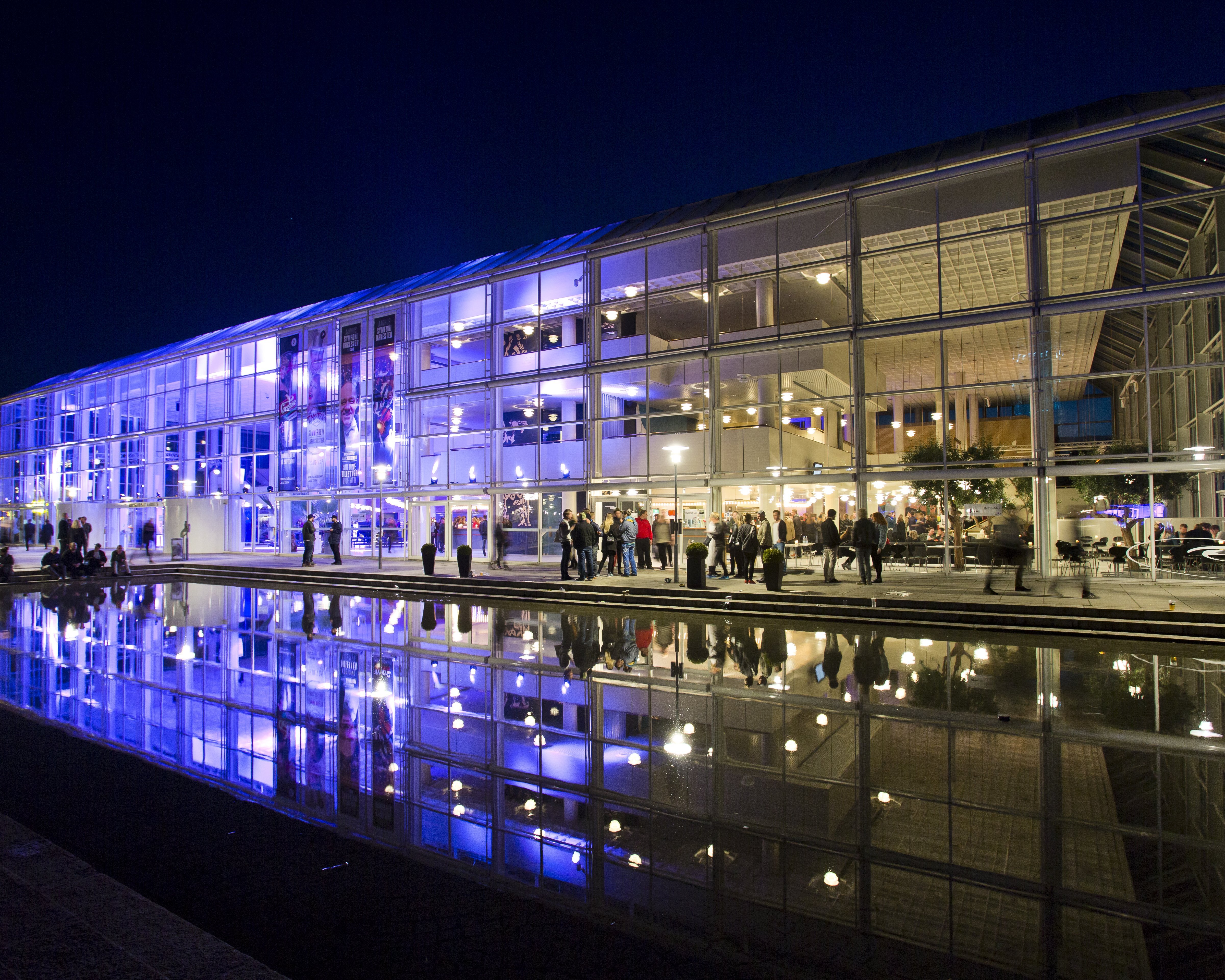 A glass building lit with blue and white lights stands at night, reflected clearly in a large pool of water in front, while people gather around the entrance.