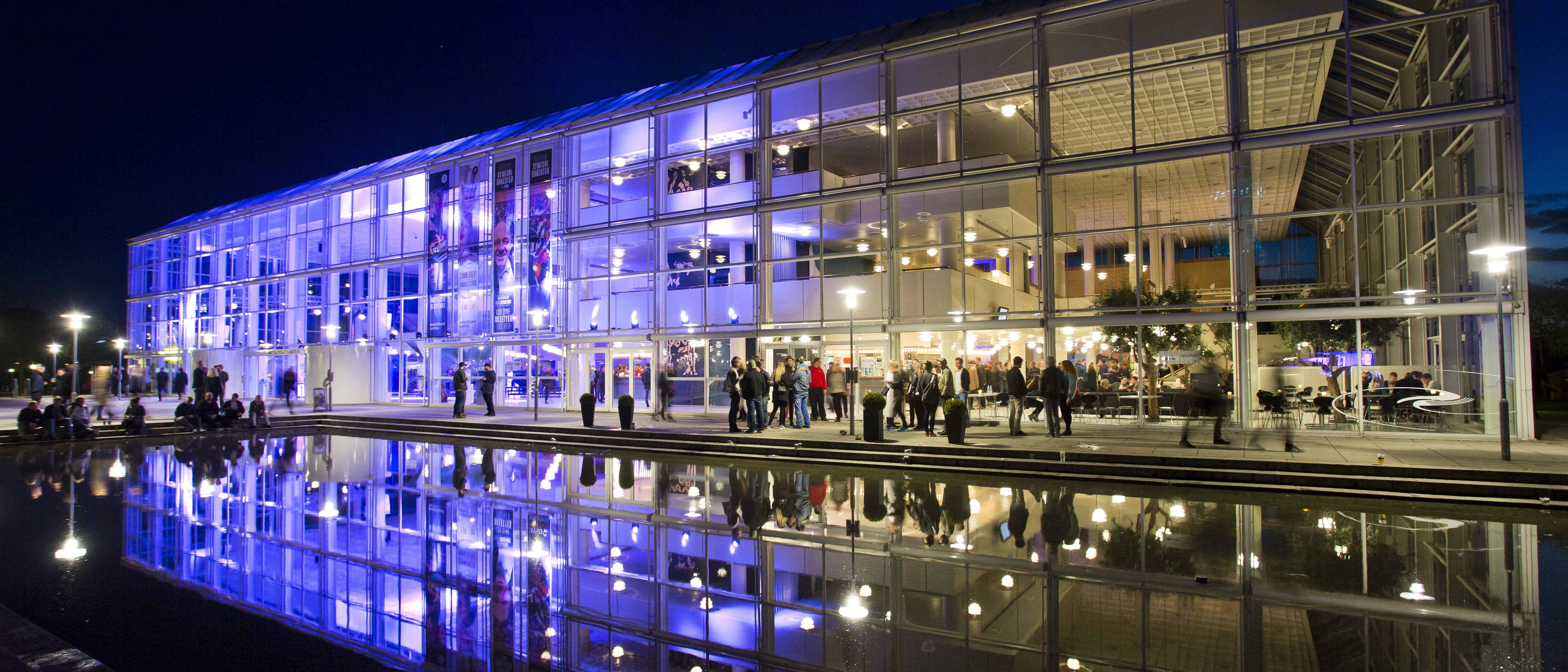 Ein modernes Glasgebäude, das nachts mit blauen und violetten Lichtern beleuchtet wird, spiegelt sich in einem stillen Wasserbecken, in dem sich Menschen vor und in dem Gebäude versammelt haben.