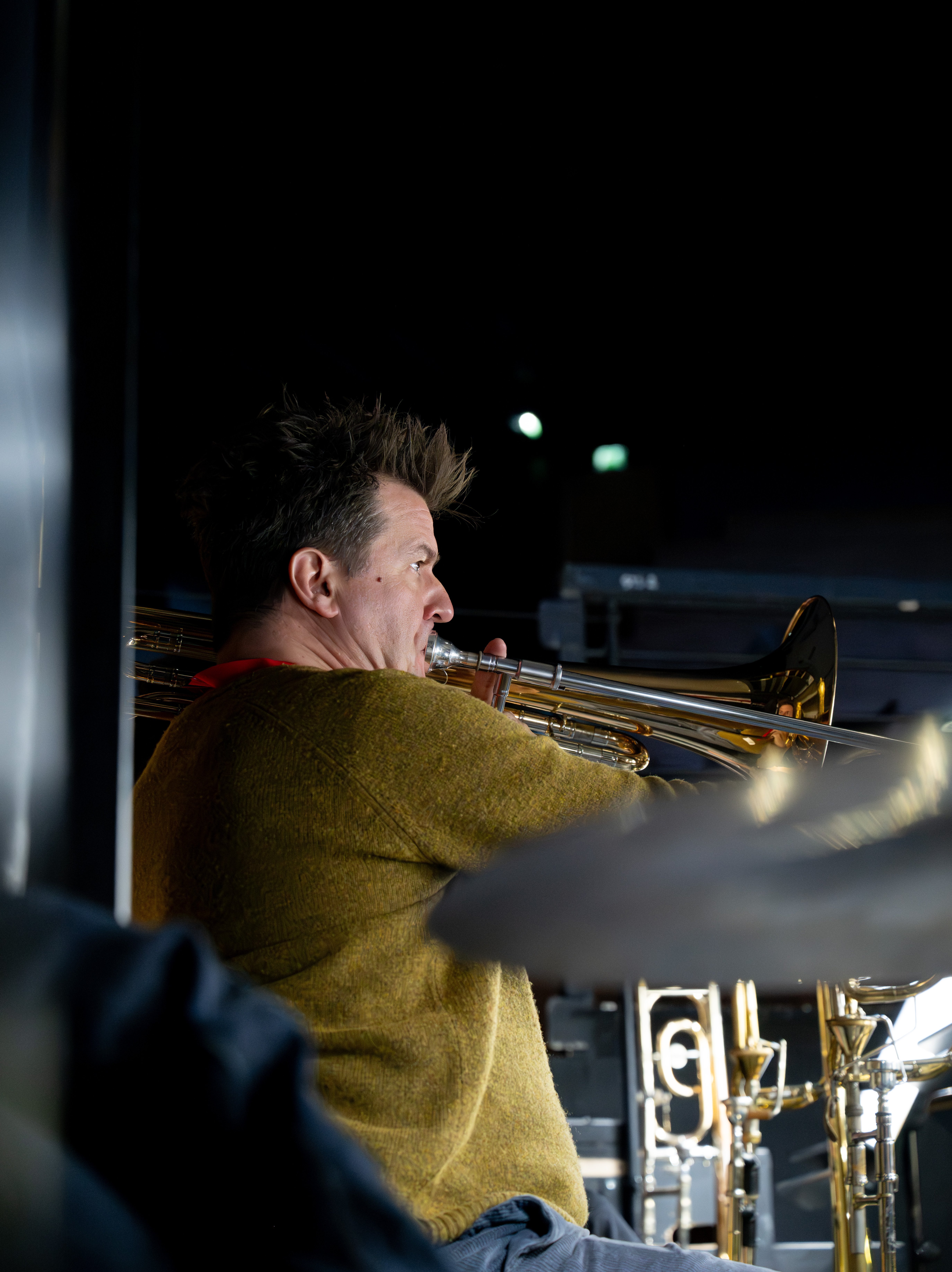 Eine Person in einem senffarbenen Pullover spielt eine Posaune, in einer schwach beleuchteten Umgebung mit Musikständern und Instrumenten um sie herum.