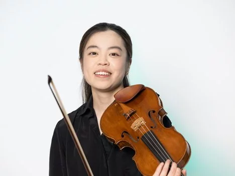 Josephine Chung with long dark hair smiles while holding a violin and bow, wearing a black top, standing against a plain white background.
