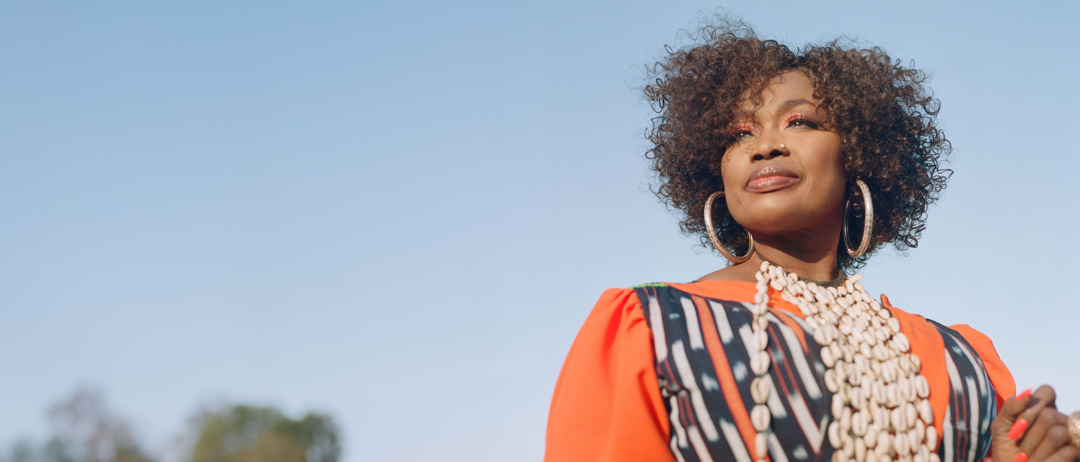 Singer Oumou Sangaré with curly hair in an orange-coloured, patterned dress and shell necklace against a blue sky, surrounded by trees.