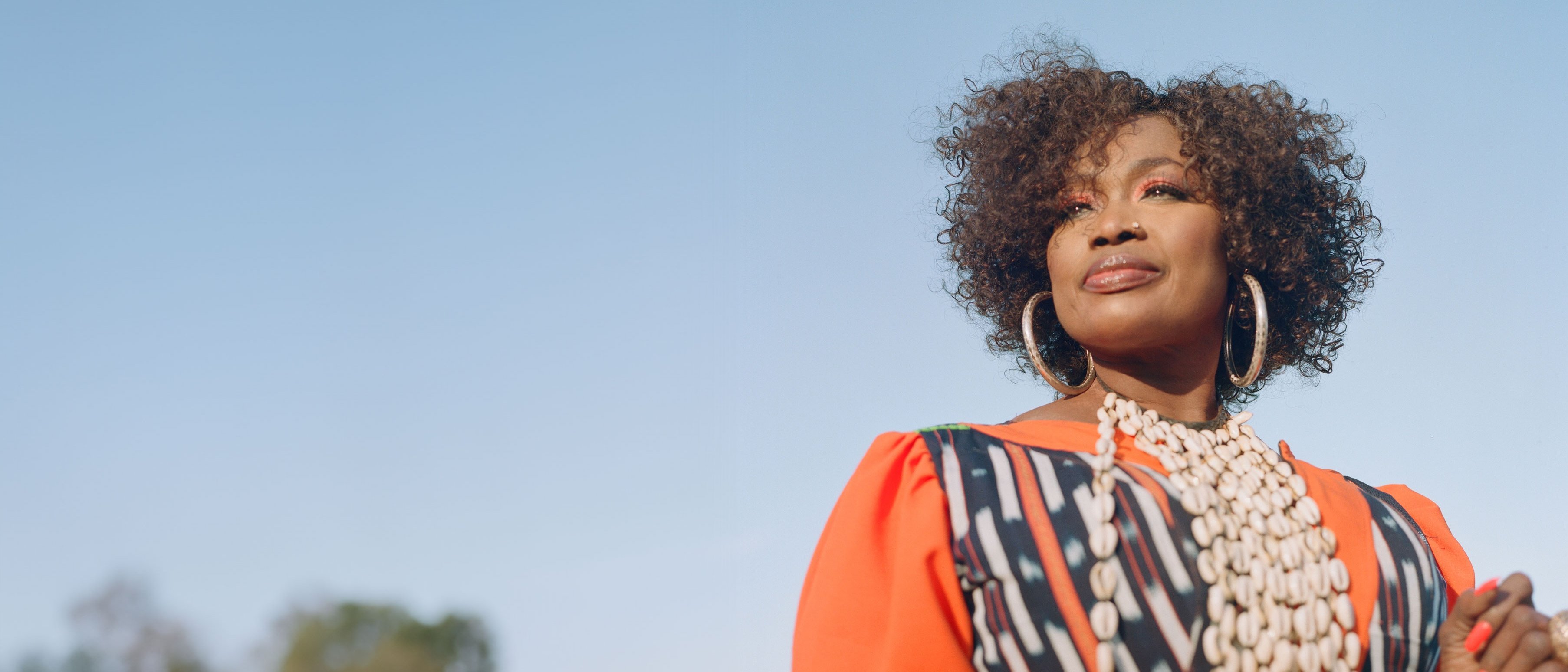 Singer Oumou Sangaré with curly hair in an orange-coloured, patterned dress and shell necklace against a blue sky, surrounded by trees.
