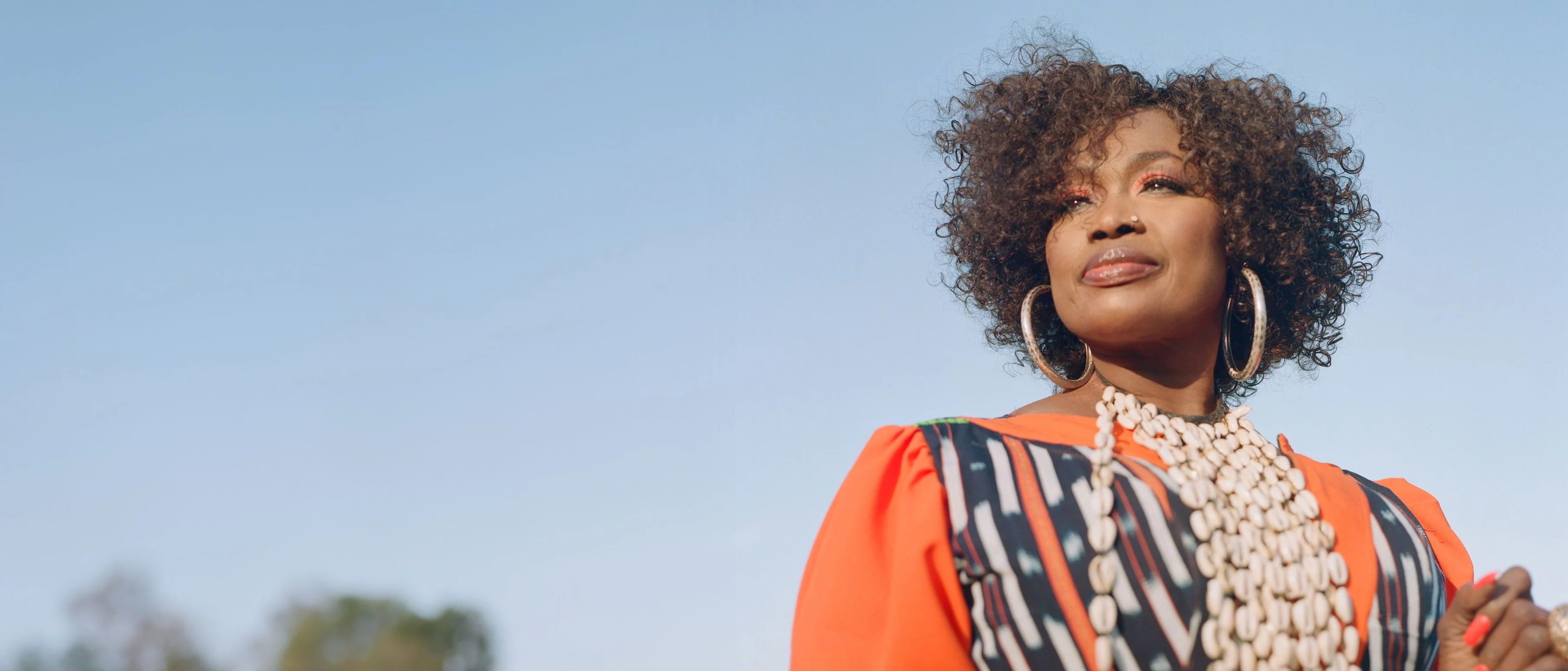 Sängerin Oumou Sangaré mit lockigem Haar in einem orangefarbenen, gemusterten Kleid und Muschelkette vor blauem Himmel, umgeben von Bäumen.
