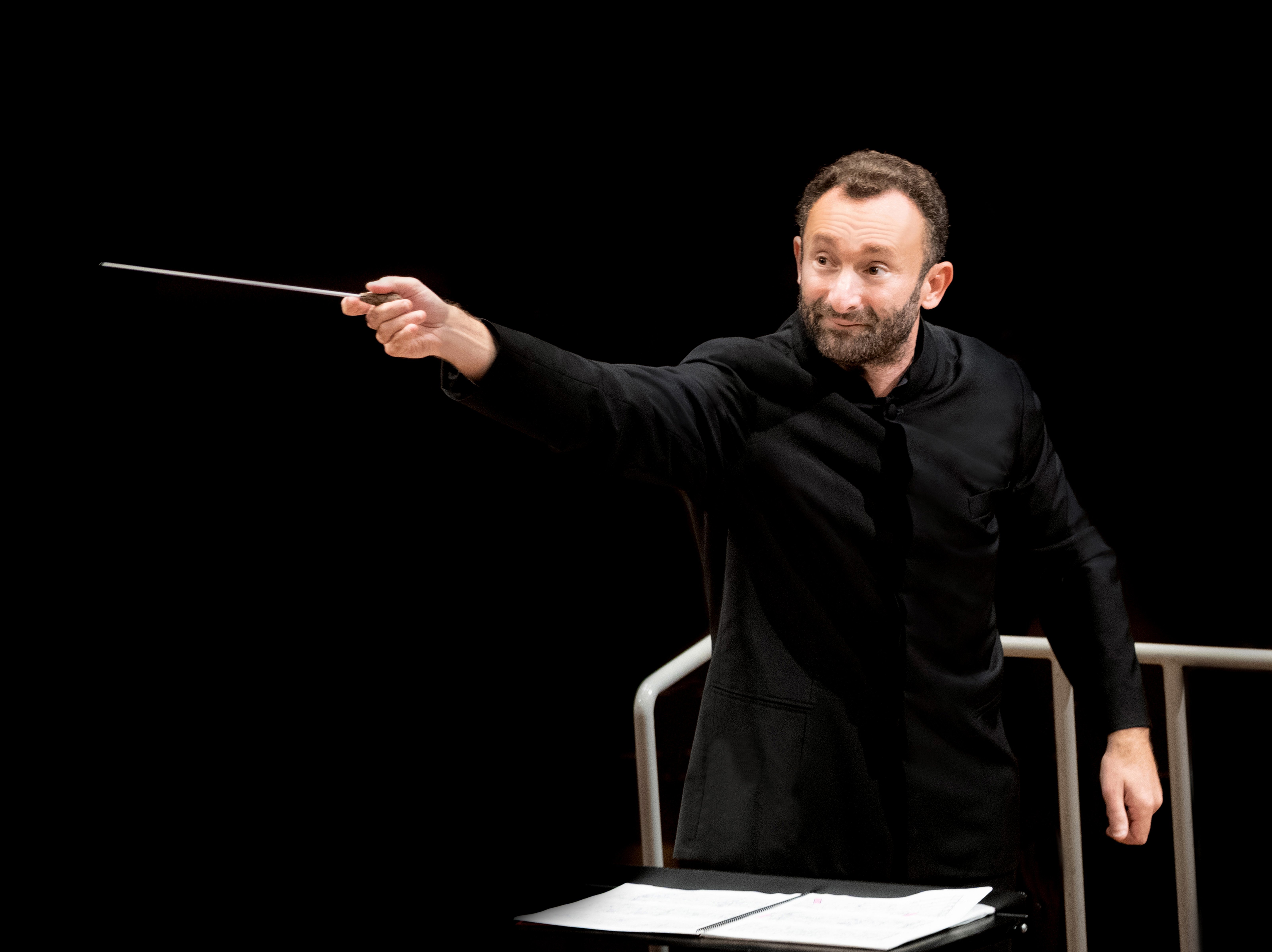 Kirill Petrenko im schwarzen Anzug dirigiert schwungvoll mit einem Taktstock das Orchester. Er steht vor einem Notenpult vor einem schwarzen Hintergrund.