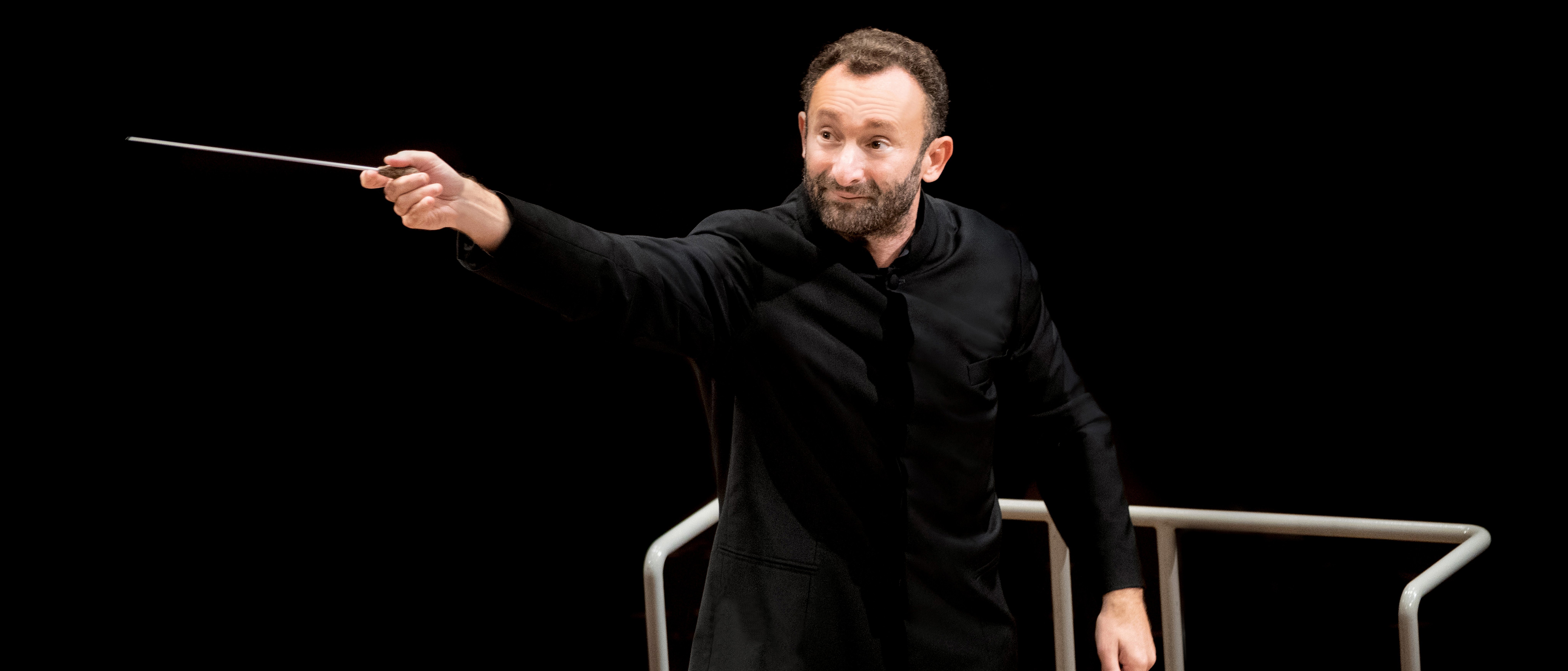 Kirill Petrenko im schwarzen Anzug dirigiert schwungvoll mit einem Taktstock das Orchester. Er steht vor einem Notenpult vor einem schwarzen Hintergrund.
