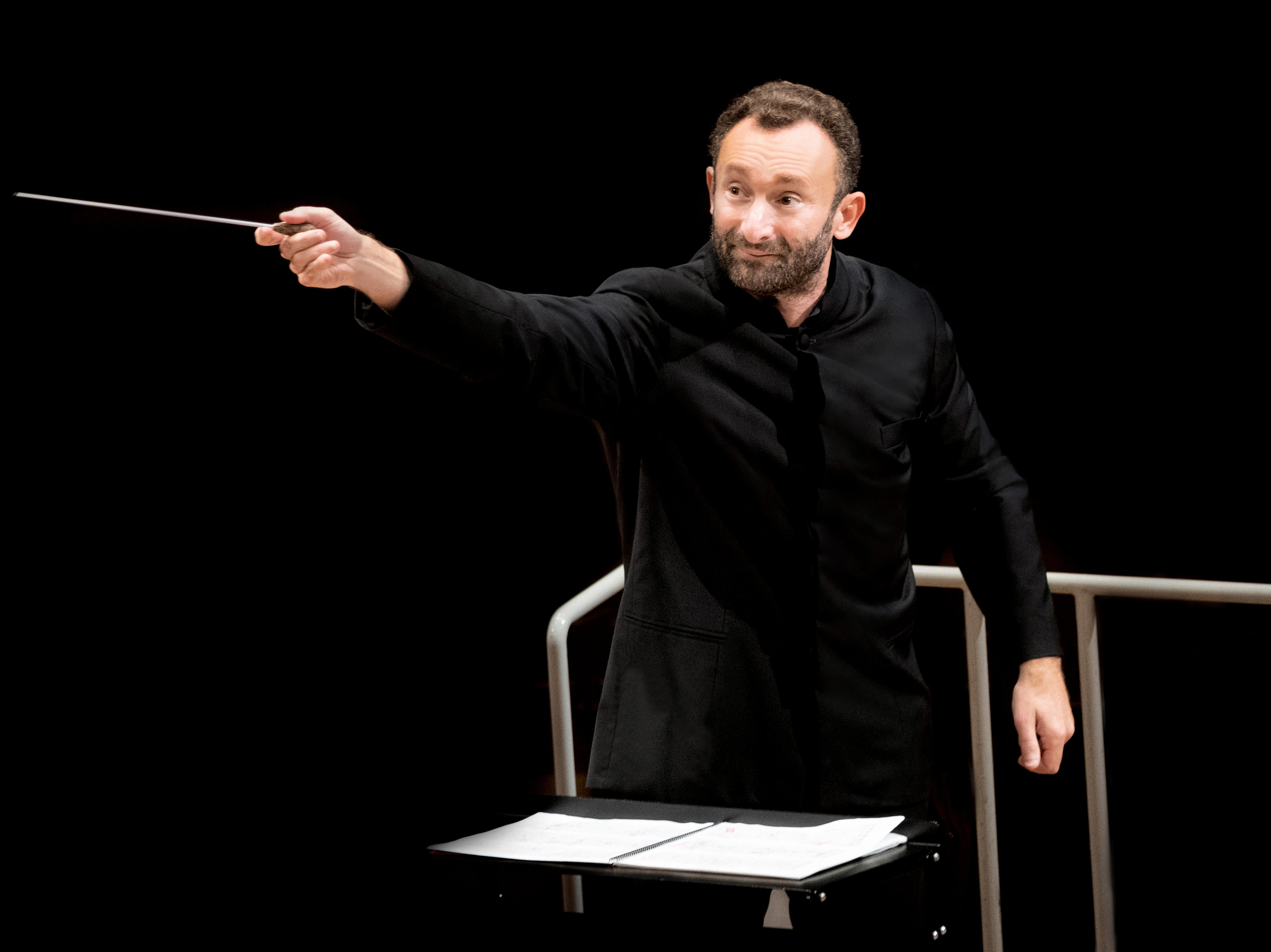 Kirill Petrenko in a black suit conducts the orchestra with a baton. He stands in front of a music stand against a black background.