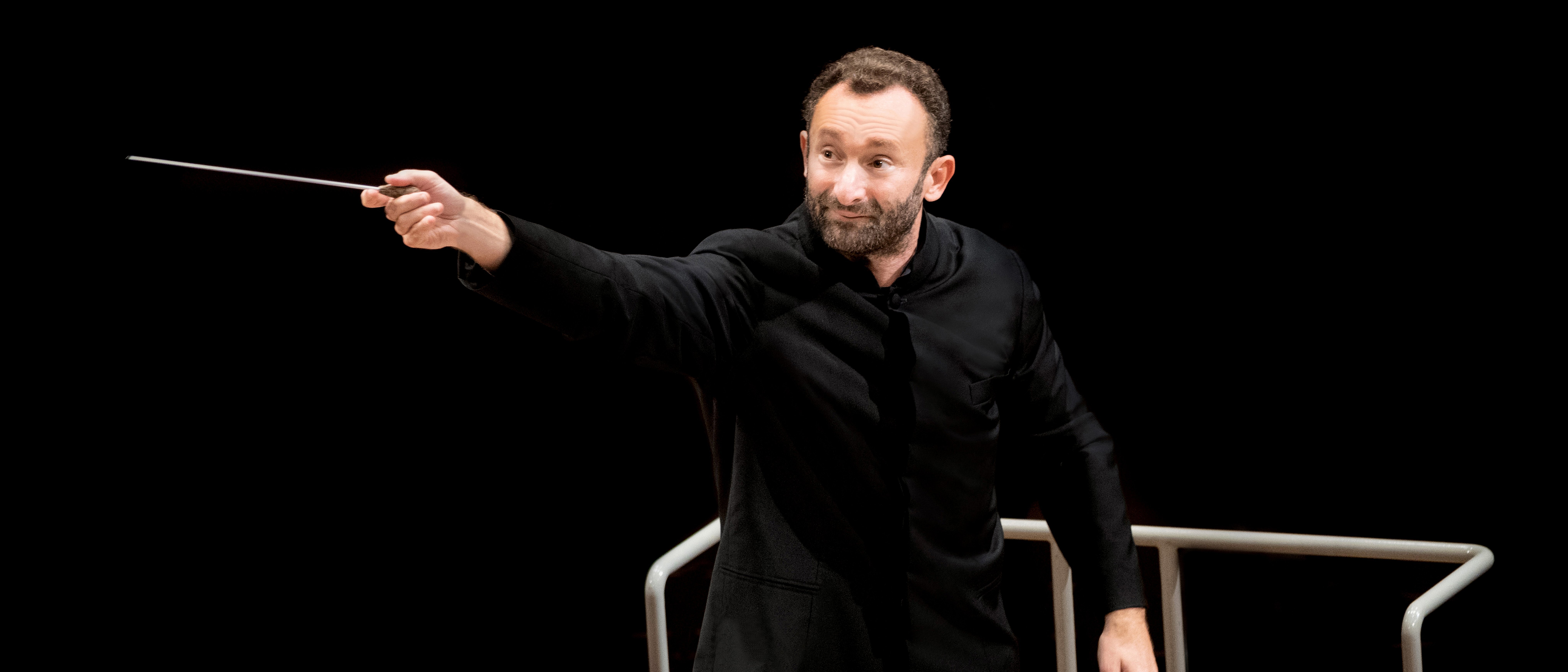 Kirill Petrenko im schwarzen Anzug dirigiert schwungvoll mit einem Taktstock das Orchester. Er steht vor einem Notenpult vor einem schwarzen Hintergrund.