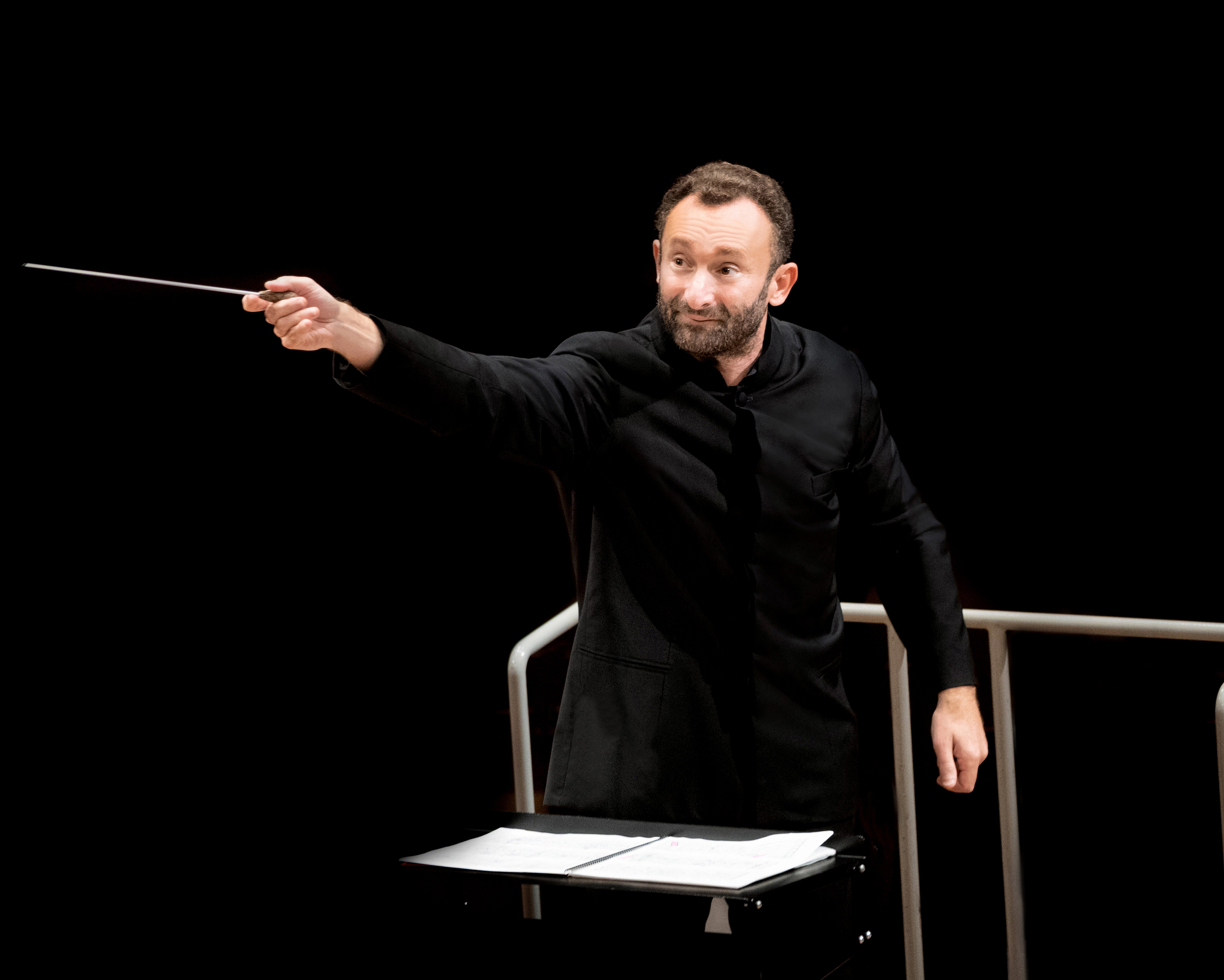 Kirill Petrenko im schwarzen Anzug dirigiert schwungvoll mit einem Taktstock das Orchester. Er steht vor einem Notenpult vor einem schwarzen Hintergrund.