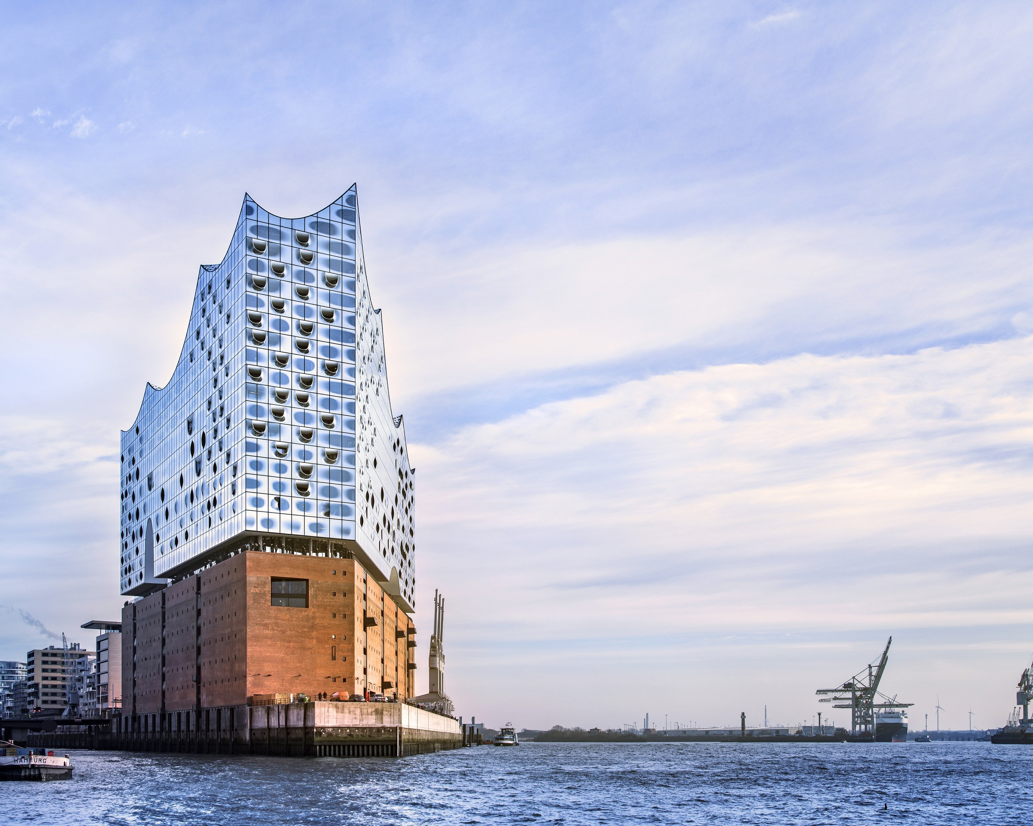 Modern glass Elbphilharmonie concert hall with a wave-shaped roof stands on a brick base by the water in Hamburg, Germany, with cranes and harbor in the background under a partly cloudy sky.