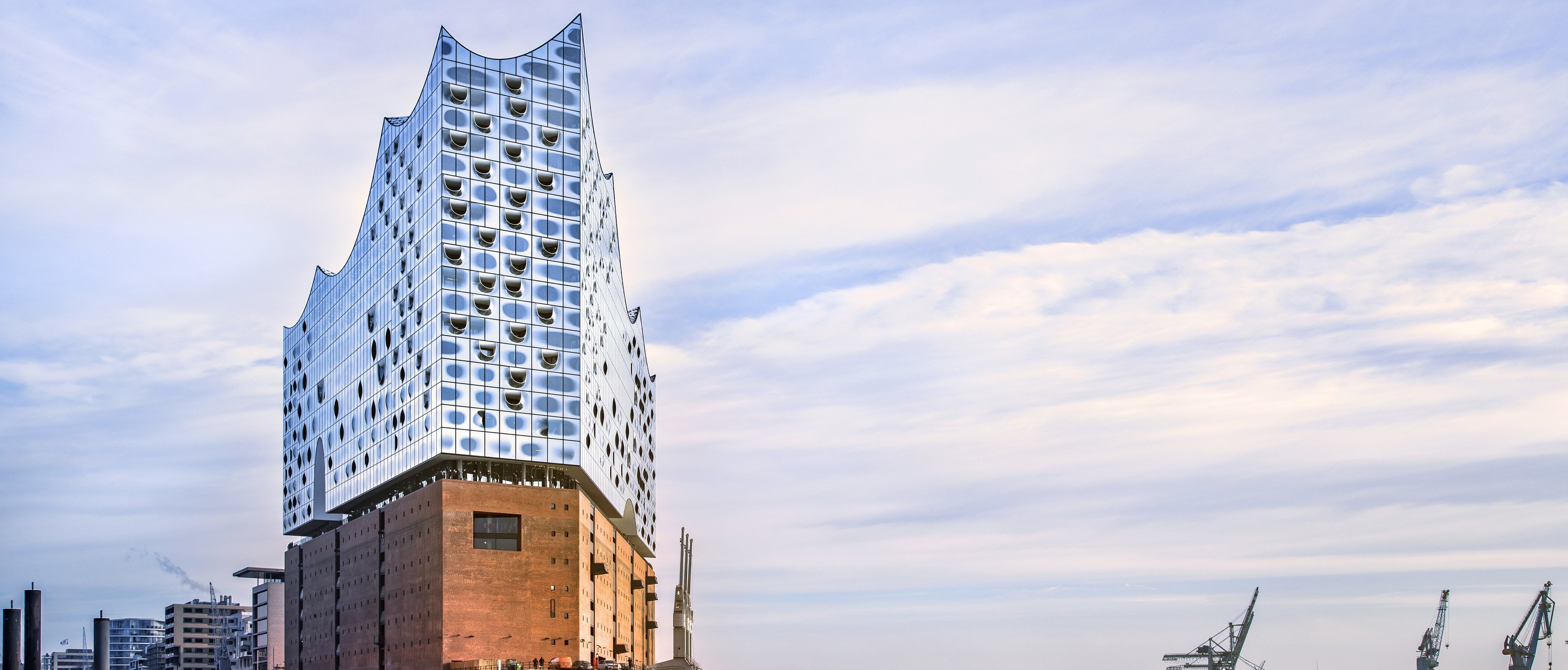 Ein modernes Glasgebäude mit einem wellenförmigen Dach steht auf einem Backsteinsockel am Wasser; in der Ferne sind unter einem teilweise bewölkten Himmel Kräne und Industrieanlagen zu sehen.
