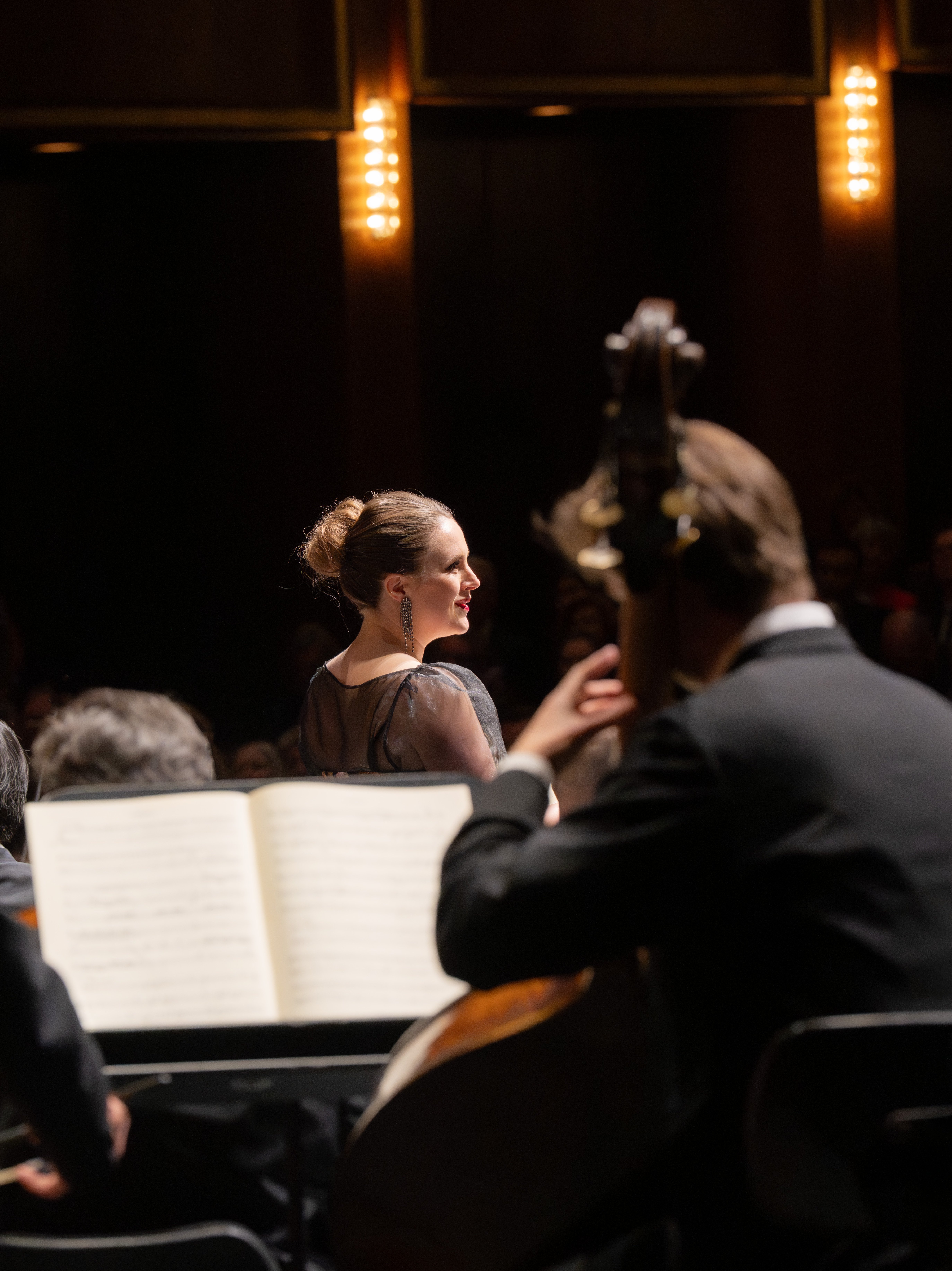 Eine Frau in einem formellen Kleid steht auf der Bühne und blickt auf ein sitzendes Orchester. Warmes Bühnenlicht beleuchtet sie, während ein Musiker im Vordergrund ein Kontrabass spielt.