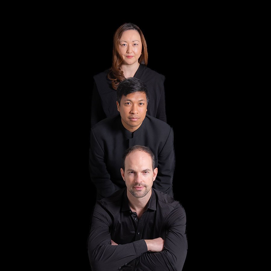 Three people in black clothing are posed in a vertical line against a black background, with a man in front, another man behind him, and a woman at the top, all facing forward.