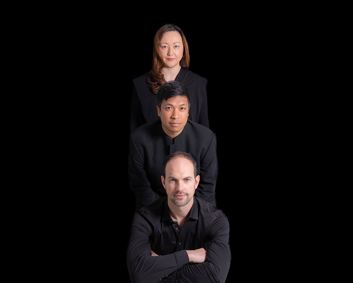 Three people in black clothing are posed in a vertical line against a black background, with a man in front, another man behind him, and a woman at the top, all facing forward.