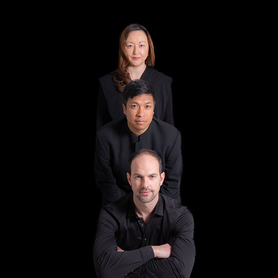 Drei Personen in schwarzer Kleidung posieren in einer vertikalen Linie vor einem schwarzen Hintergrund, mit einem Mann vorne, einem Mann in der Mitte und einer Frau hinten, alle blicken nach vorne.