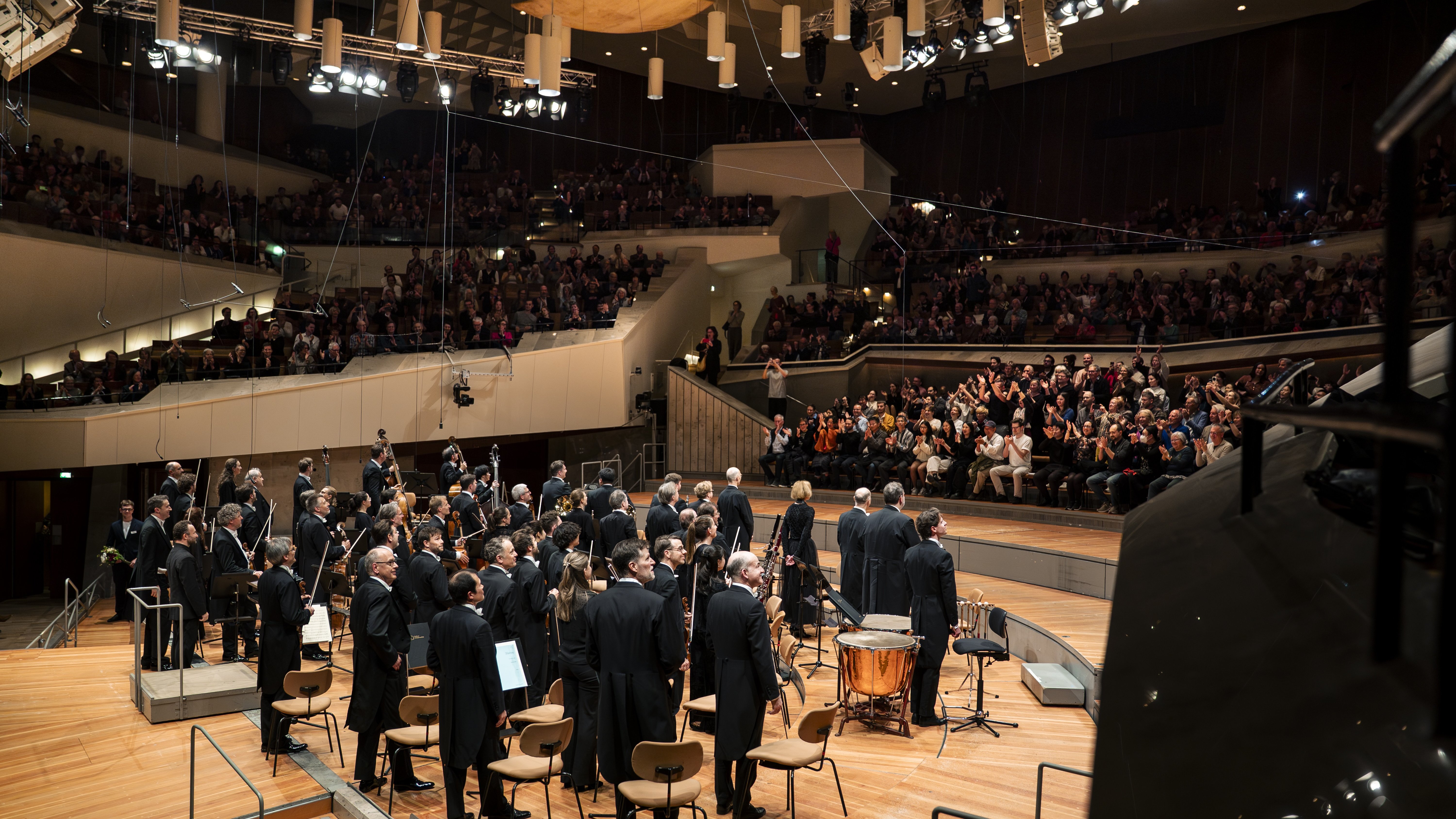 Ein Orchester steht auf der Bühne vor einem großen Publikum in einem modernen Konzertsaal. Das Publikum sitzt in Reihen und spendet den Musikern herzlichen Applaus. Der Saal ist durch Deckenbeleuchtung hell ausgeleuchtet.